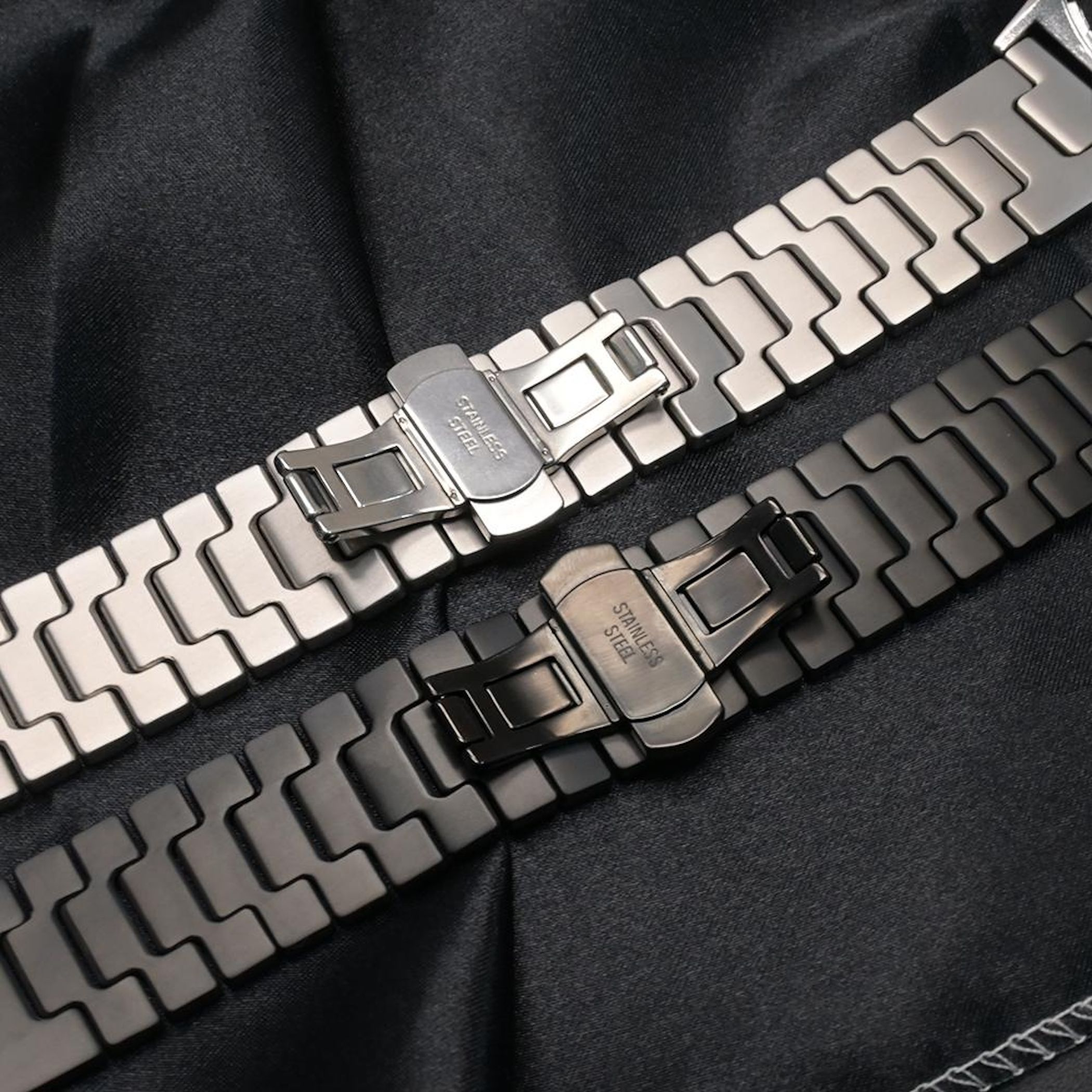 Zwei Edelstahl-Uhrenarmbänder, eines silber, eines dunkelgrau, auf schwarzem Stoff.