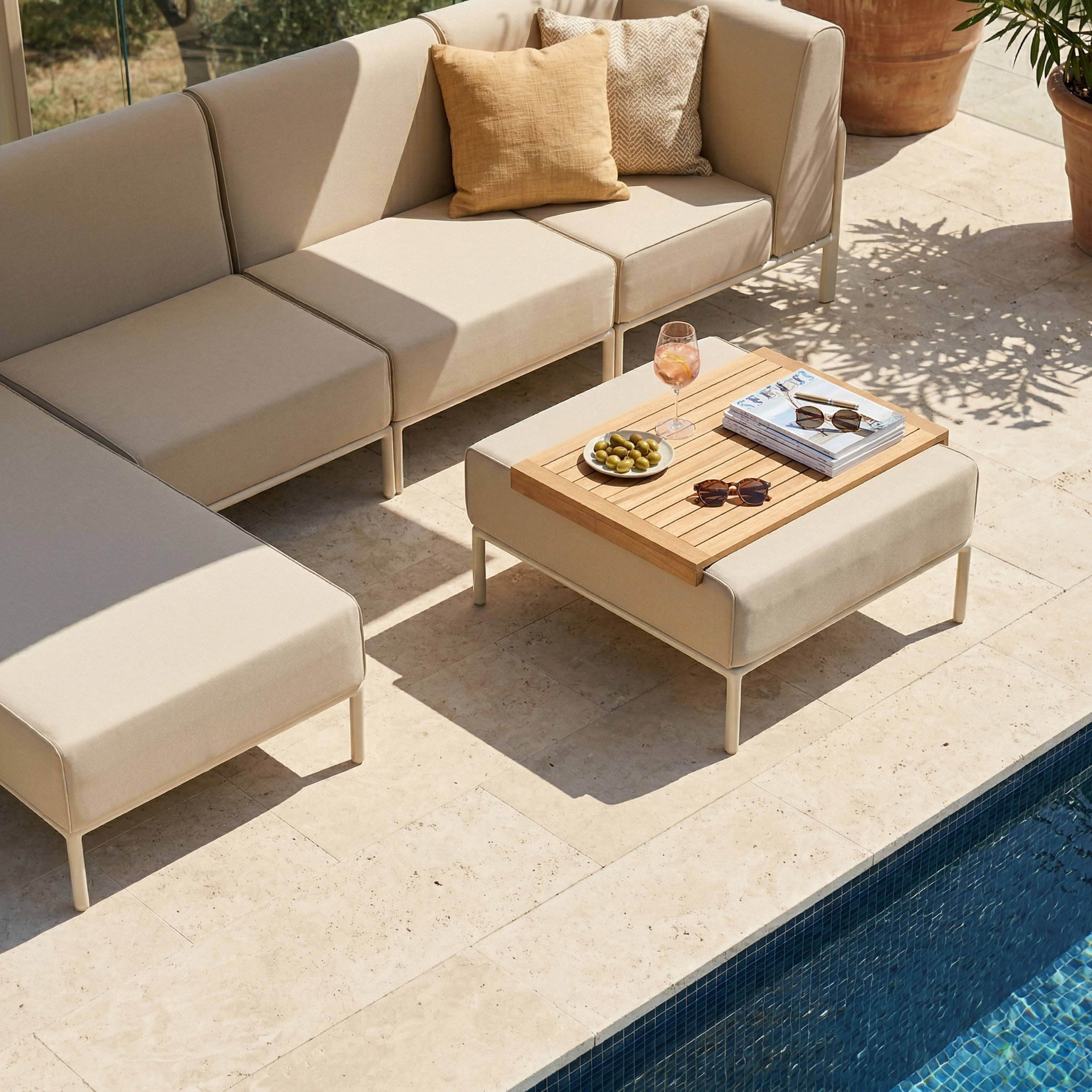 Eine beigefarbene Lounge-Garnitur mit Kissen, Couchtisch mit Getränk und Snacks am Pool.