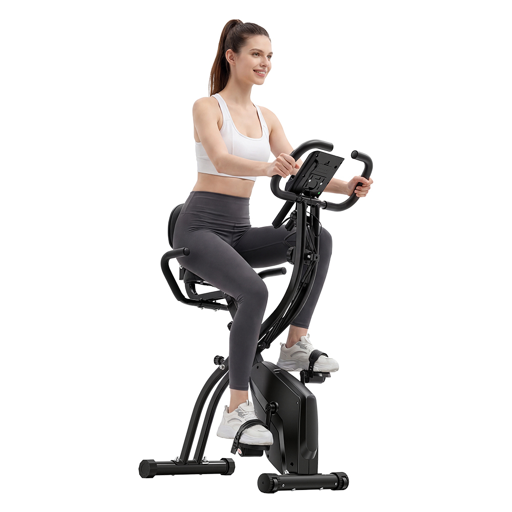 Kobieta w kucyku w białej bluzce i szarych legginsach, jeździ na rowerze stacjonarnym.