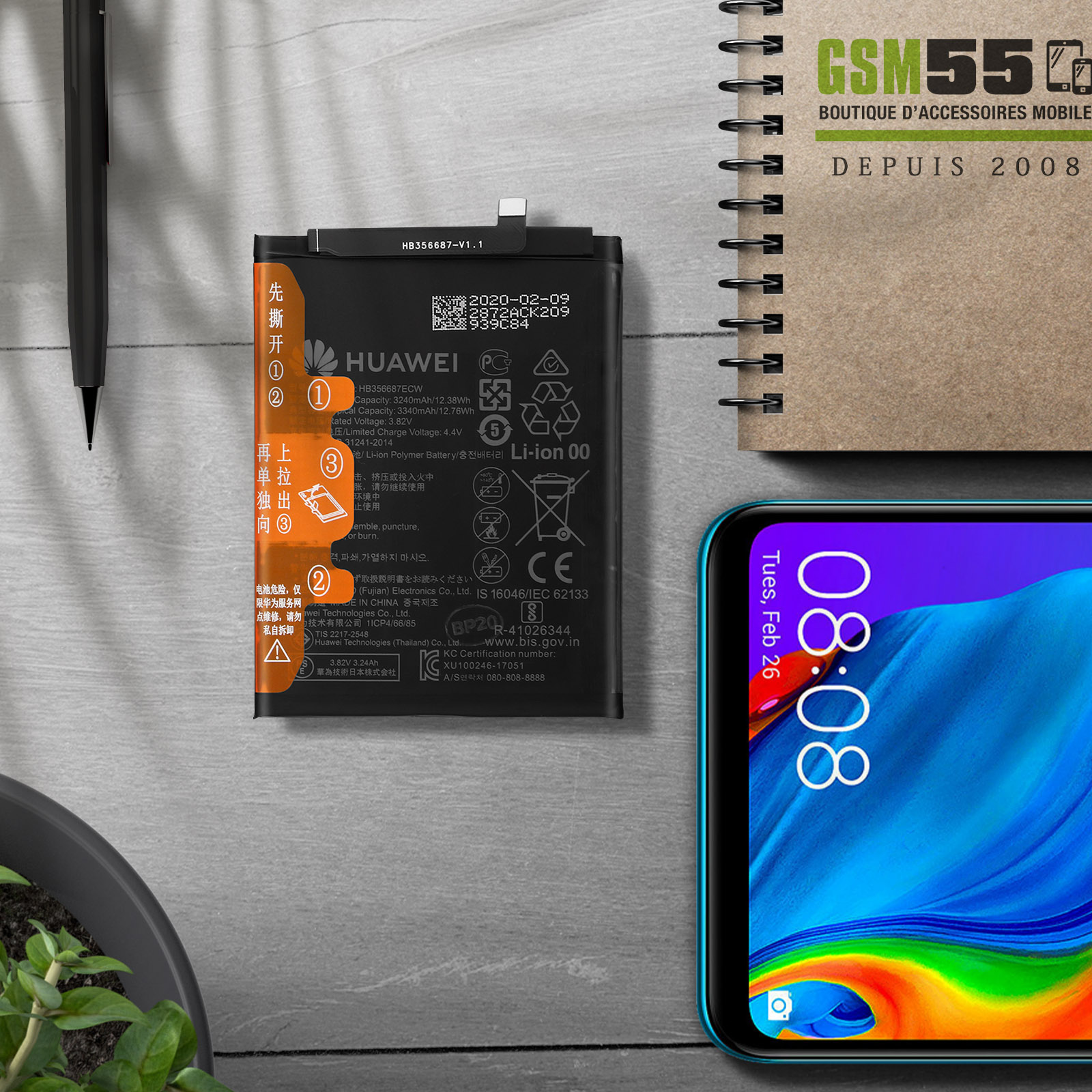 Una batteria Huawei giace su un tavolo di legno accanto a uno smartphone e un taccuino.