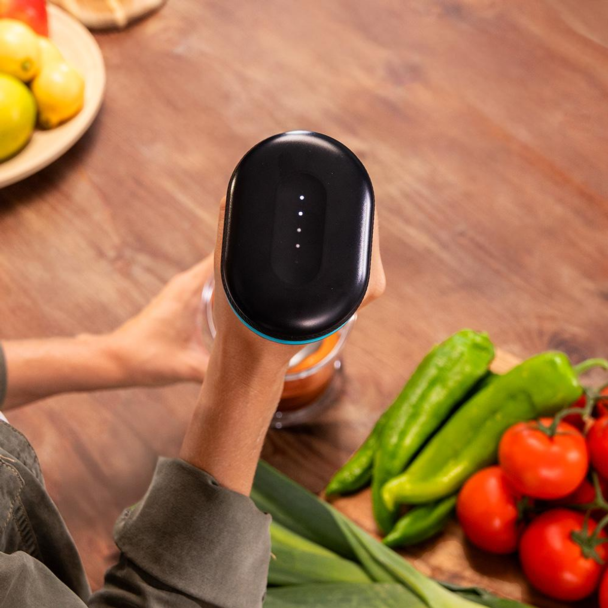 Mano sosteniendo licuadora negra sobre verduras como pimientos, tomates y puerros.