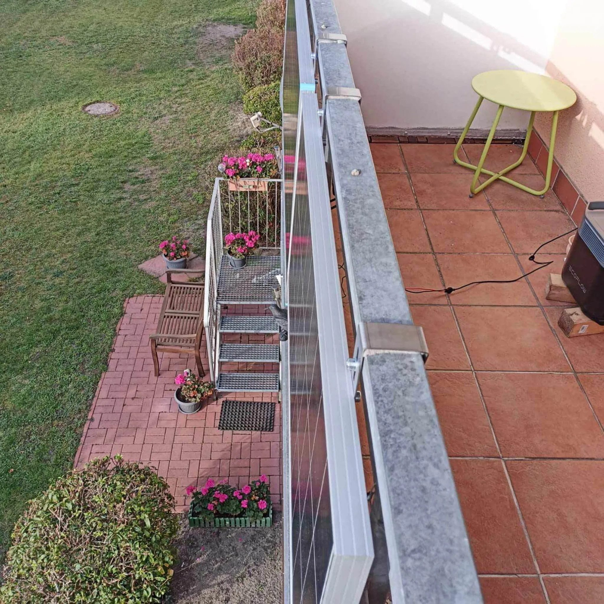 Balkon mit Solarmodulen, Metallgeländer, Sitzbank, Blumen und Tisch.