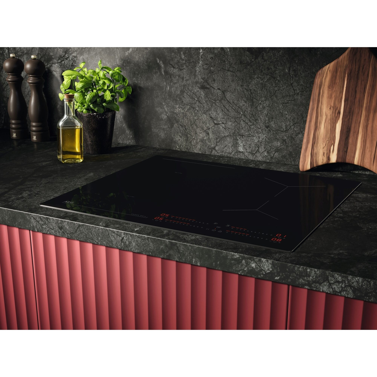 Placa de inducción negra con controles rojos, junto a aceitera, hierbas y molinillos.
