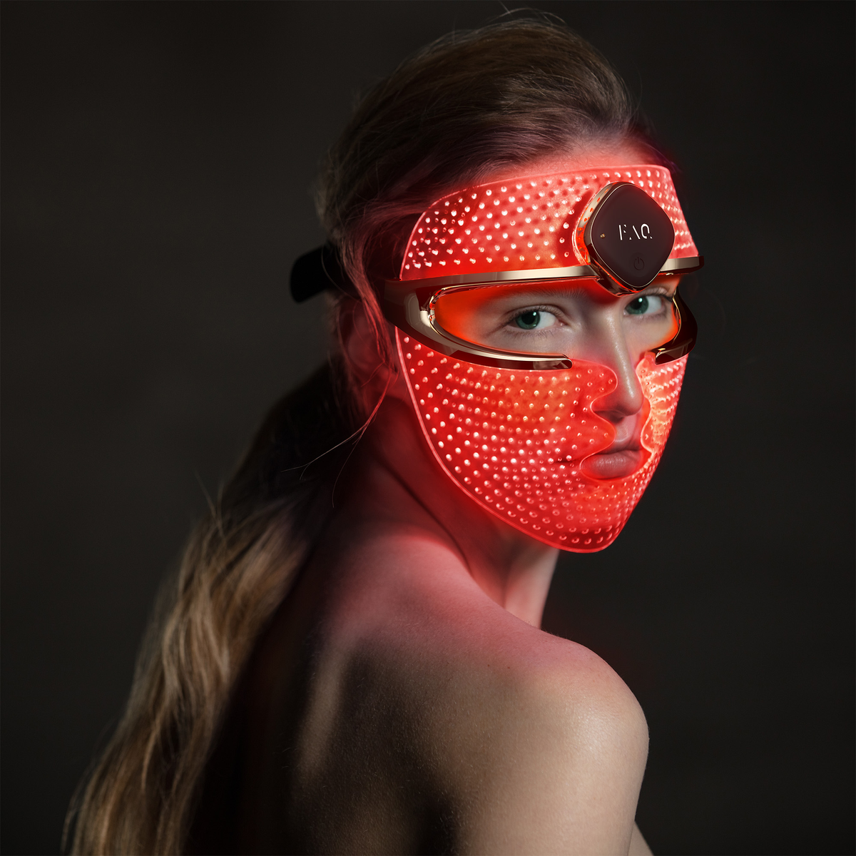 Woman wearing a glowing red LED mask with a gold frame, dark background.