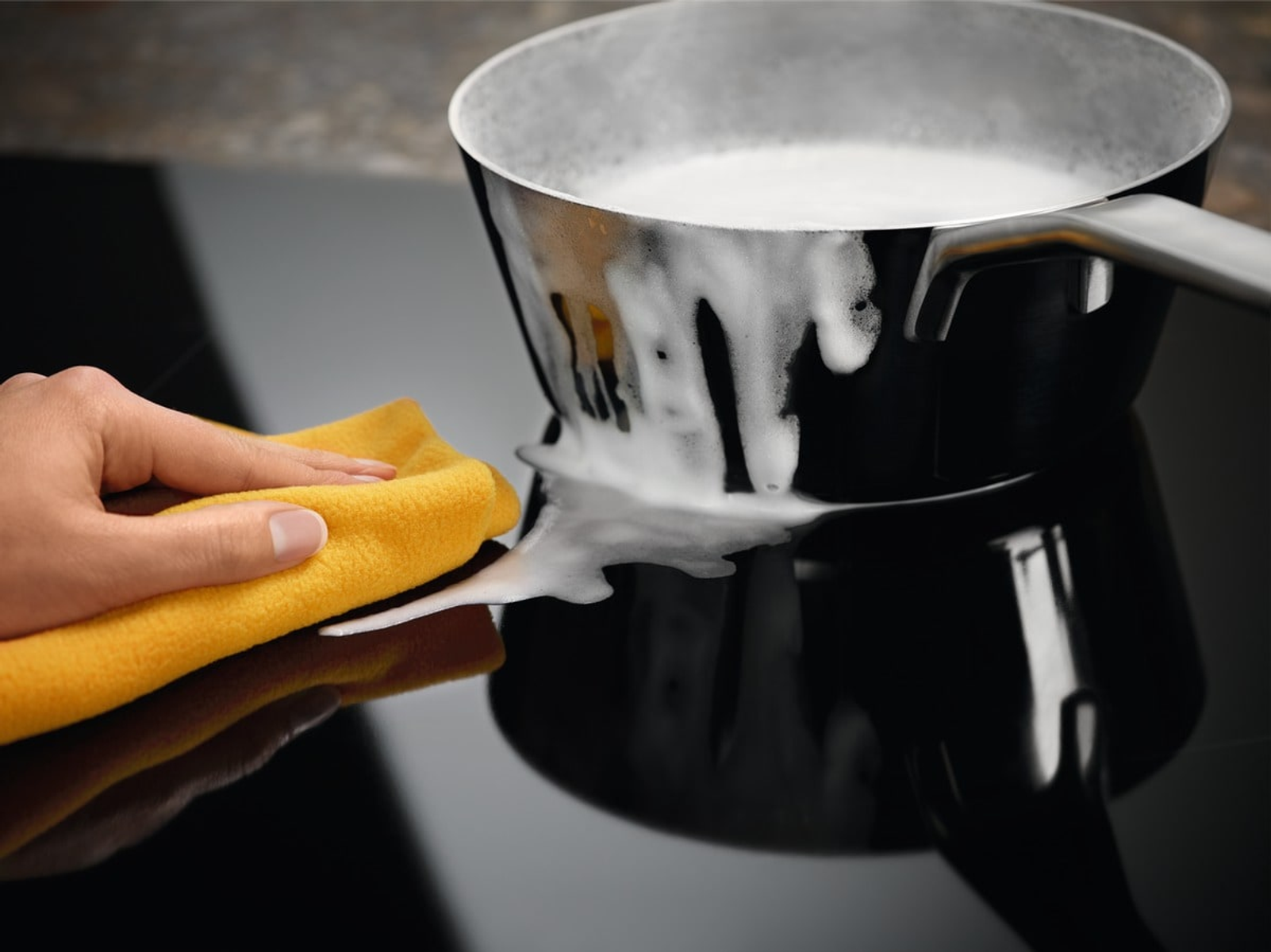 Mano con panno giallo pulisce pentola con schiuma su piano cottura.