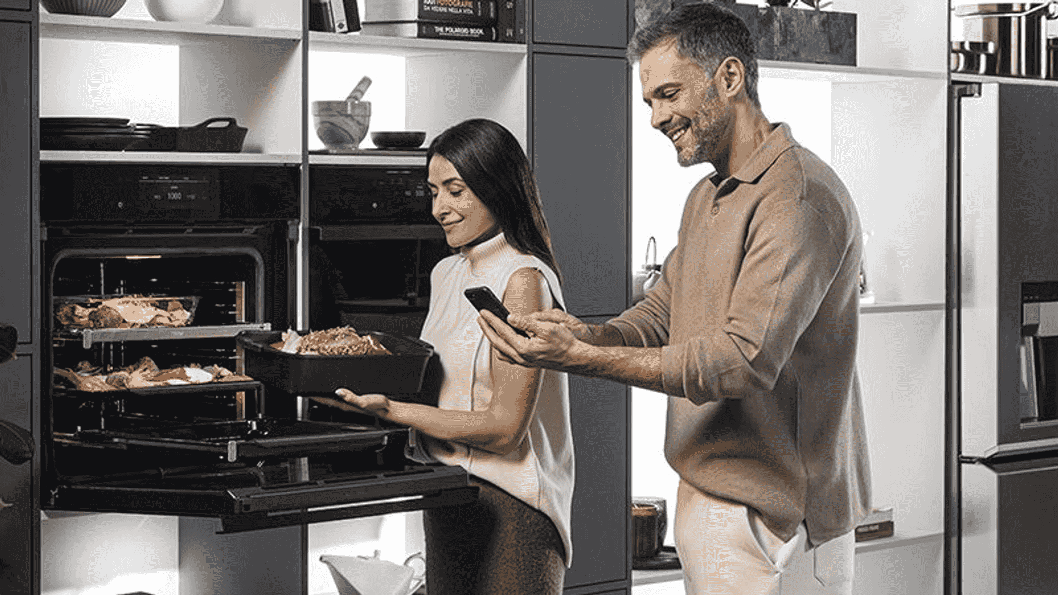 Hombre y mujer en cocina con horno. Ella sostiene una bandeja con comida, él mira su móvil.