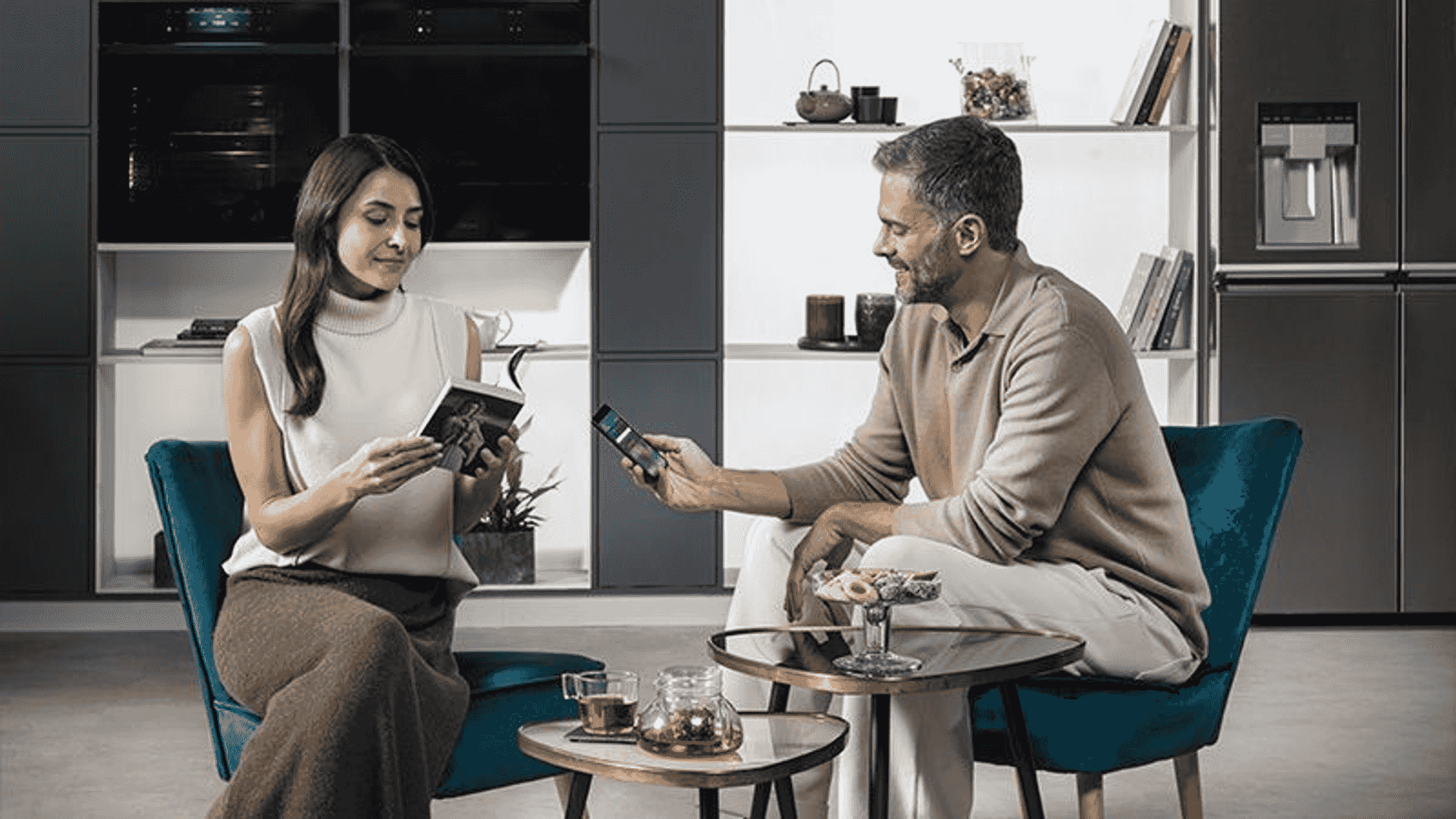 Hombre y mujer sentados en sillones, mujer lee libro, hombre mira el móvil.