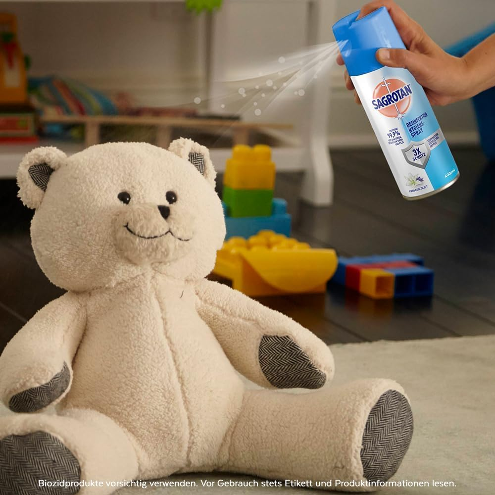 Ein Teddybär sitzt vor Spielzeug, während eine Hand Sagrotan-Spray sprüht.
