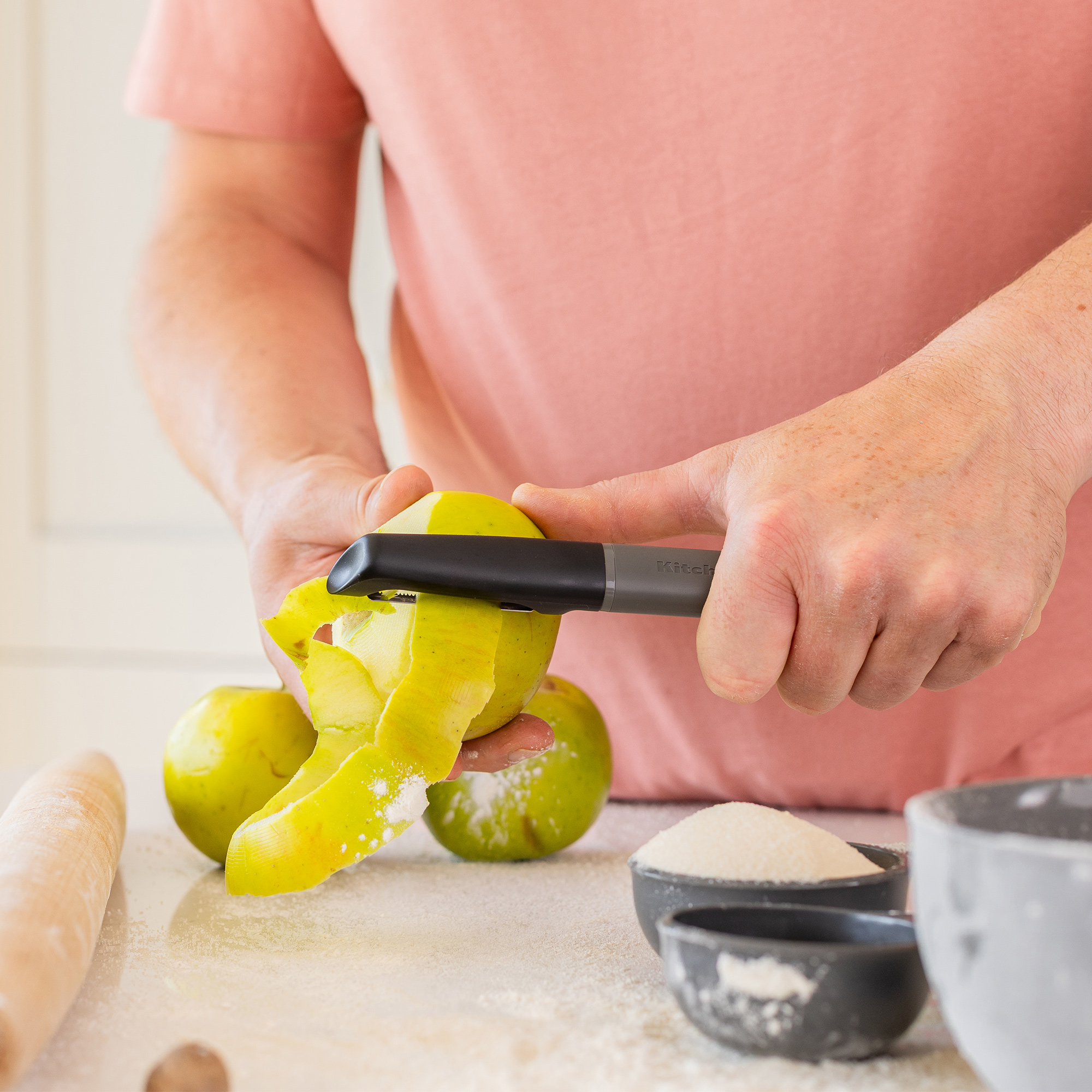 Eine Person schält einen grünen Apfel mit einem Sparschäler. Zutaten sind verstreut.