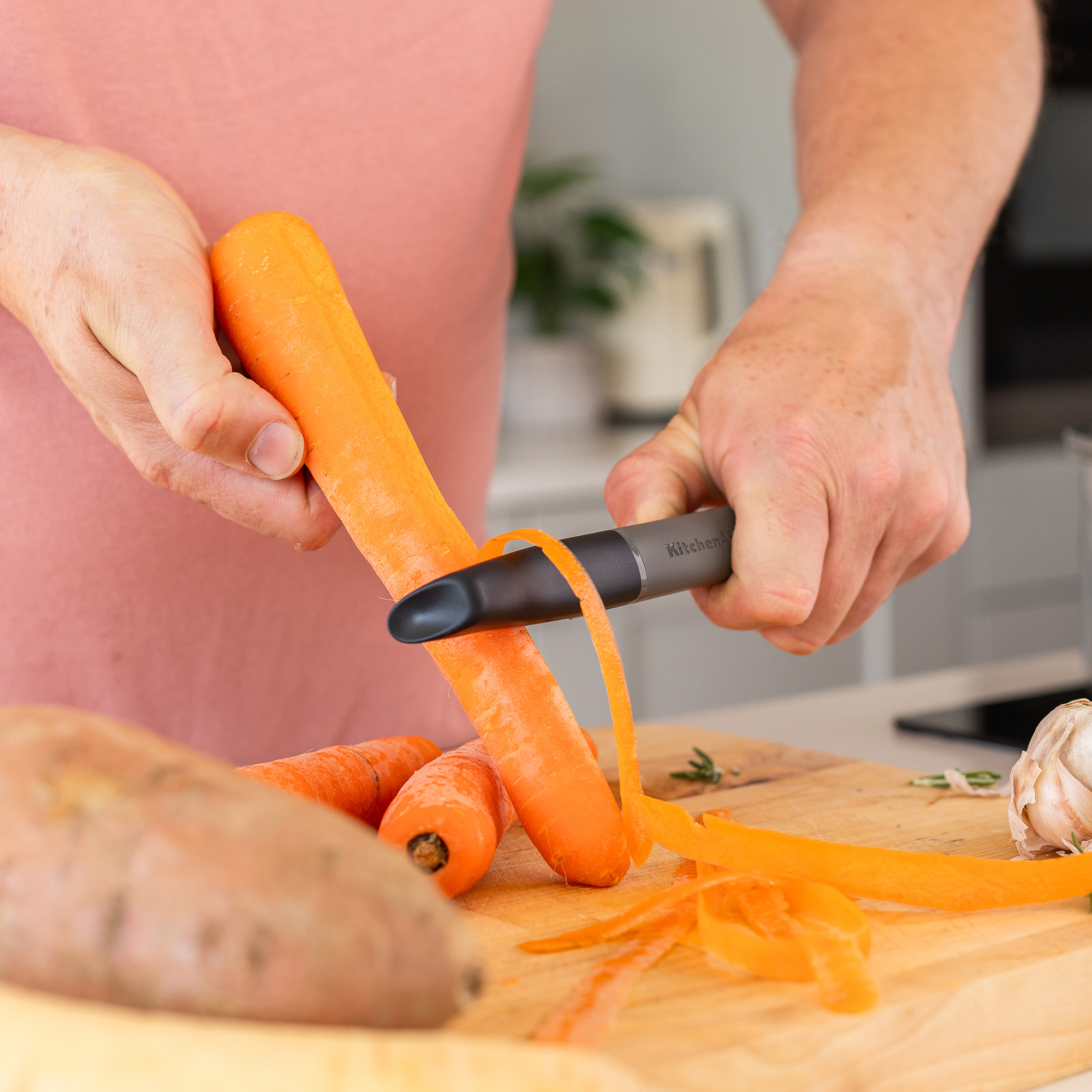 Person schält Karotten mit einem Sparschäler auf einem Holzbrett. Knoblauch und Süßkartoffel daneben.