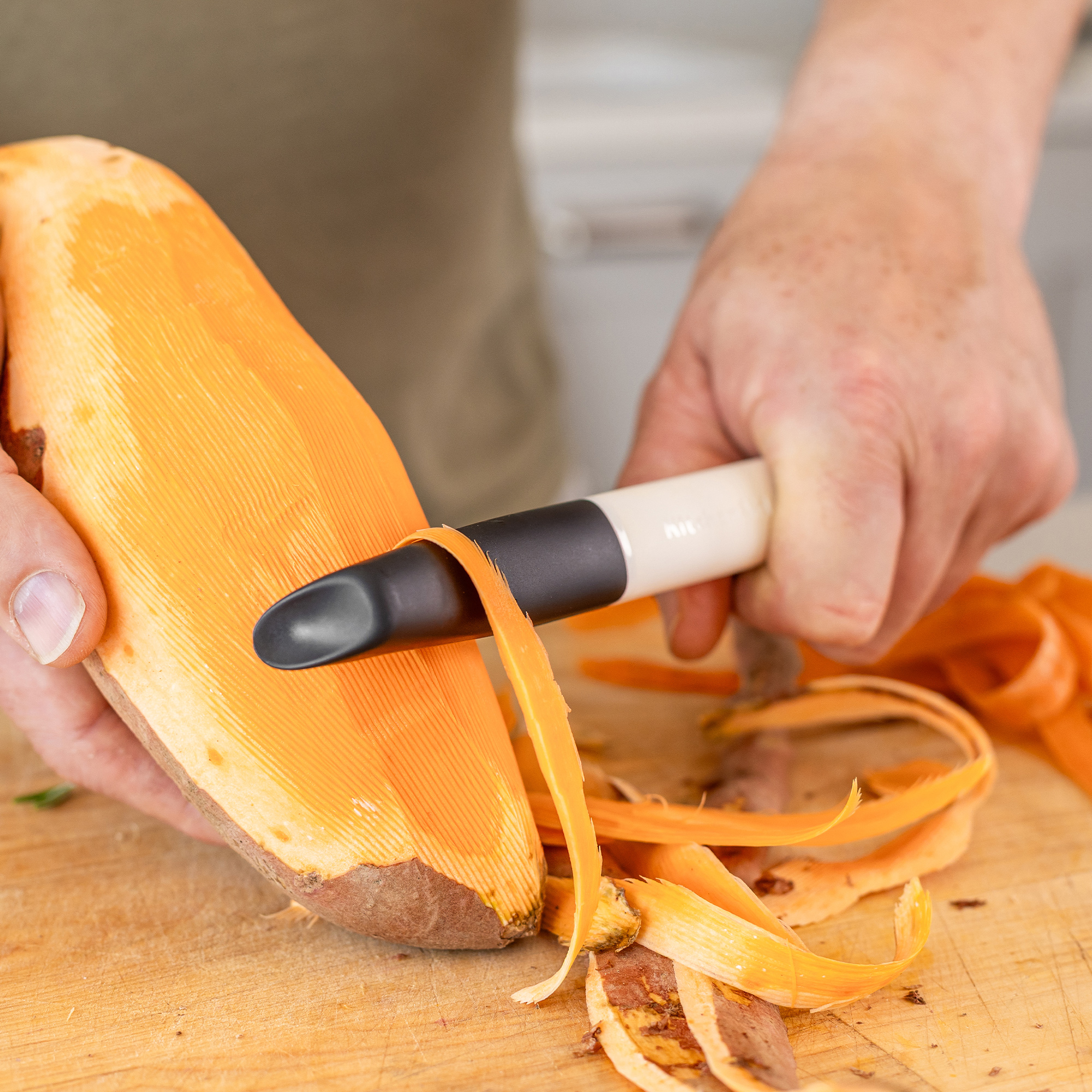Hände schälen eine Süßkartoffel auf einem Holzbrett mit einem Sparschäler.