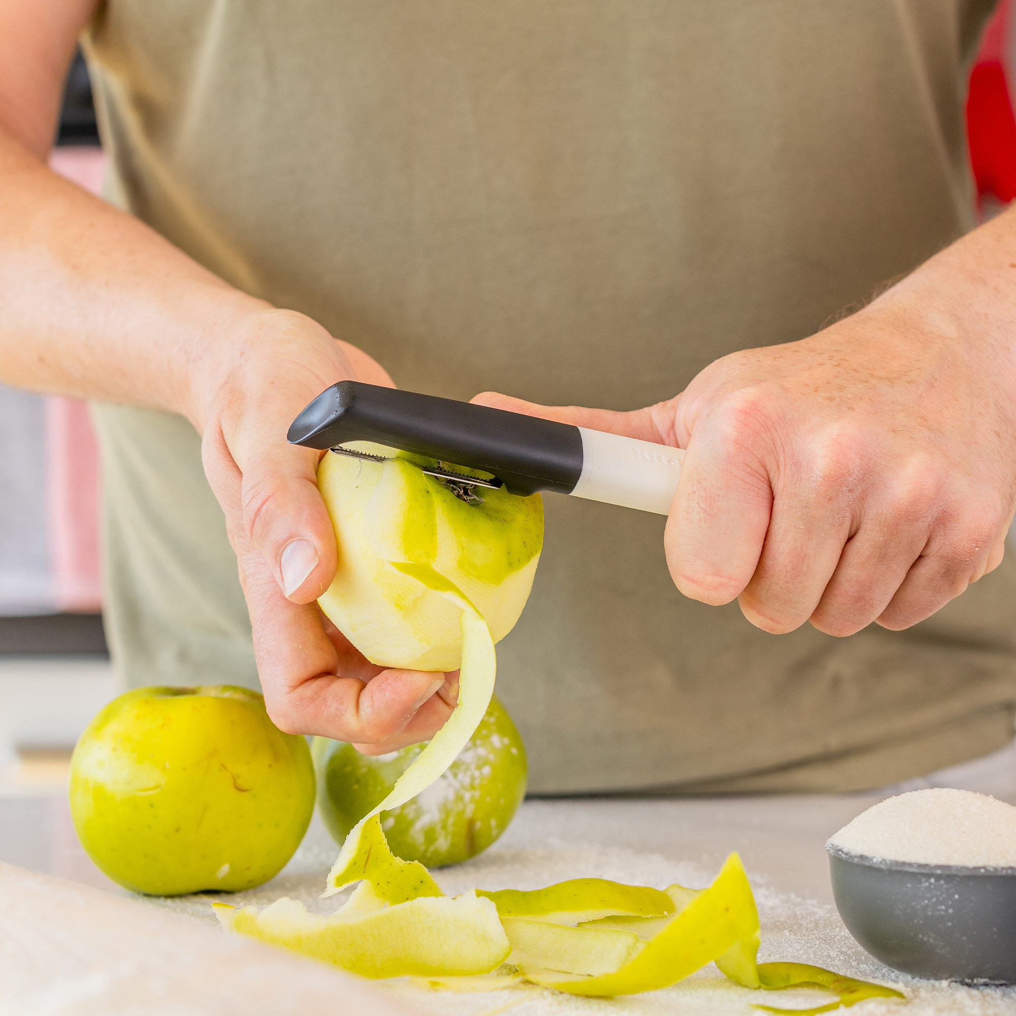 Jemand schält einen grünen Apfel mit einem Sparschäler, daneben liegen Äpfel und Zucker.