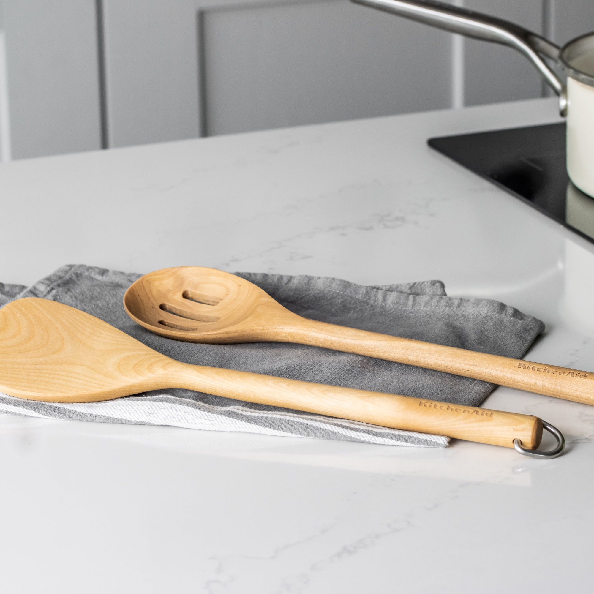 Zwei hölzerne KitchenAid-Utensilien liegen auf einem gestreiften Tuch auf einer weißen Arbeitsplatte.