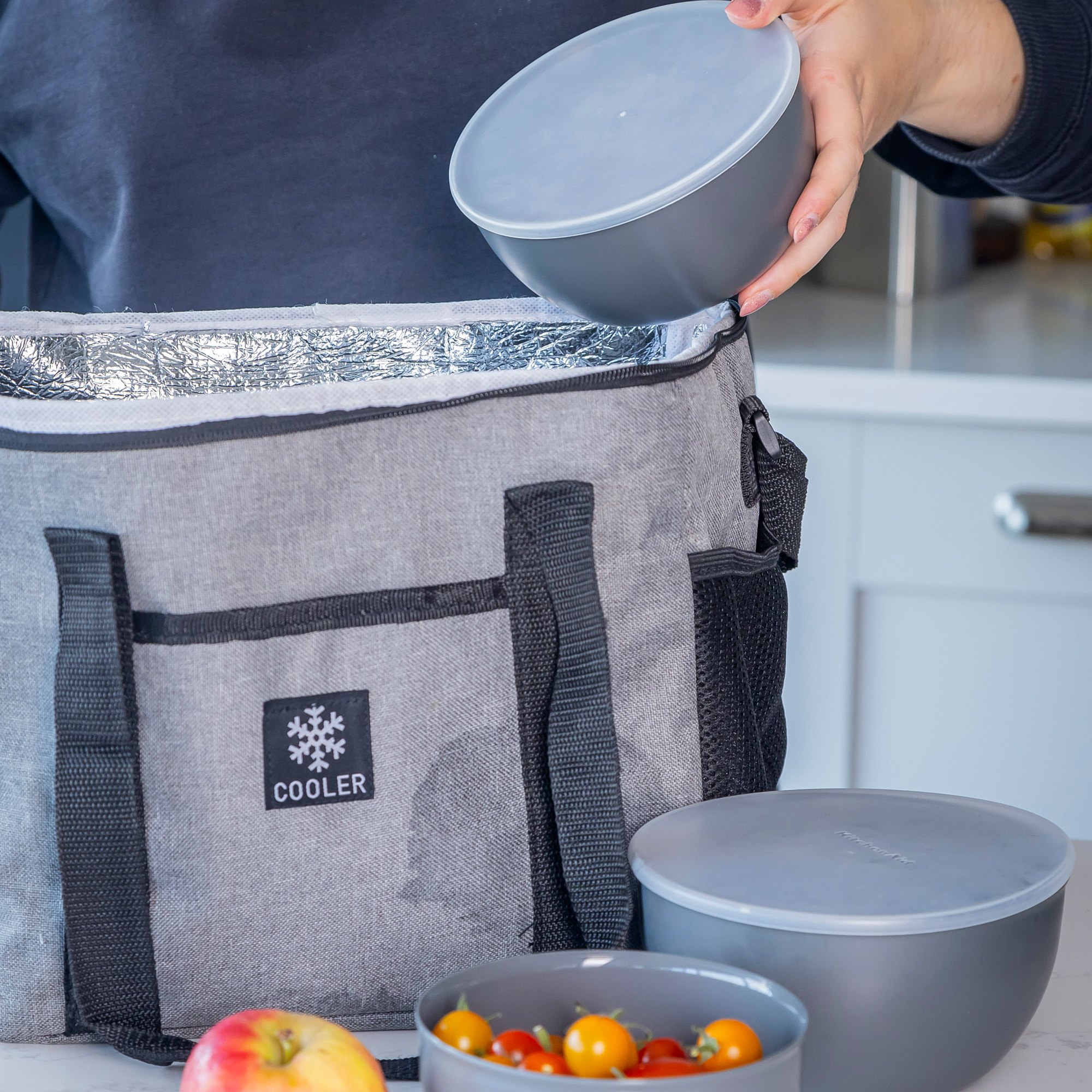 Eine Person packt eine Schüssel in eine graue Kühltasche. Daneben liegen eine Schüssel mit Tomaten und ein Apfel.