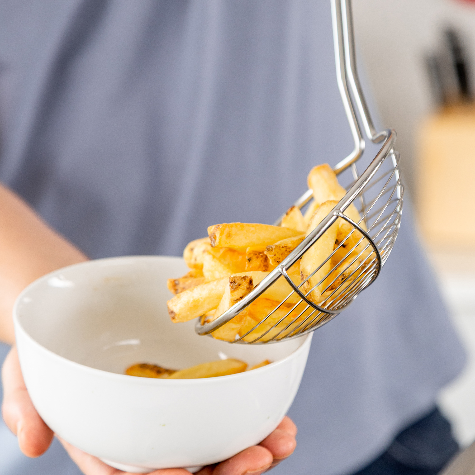 Goldbraune Pommes werden mit einem Sieb in eine weiße Schüssel gegeben.