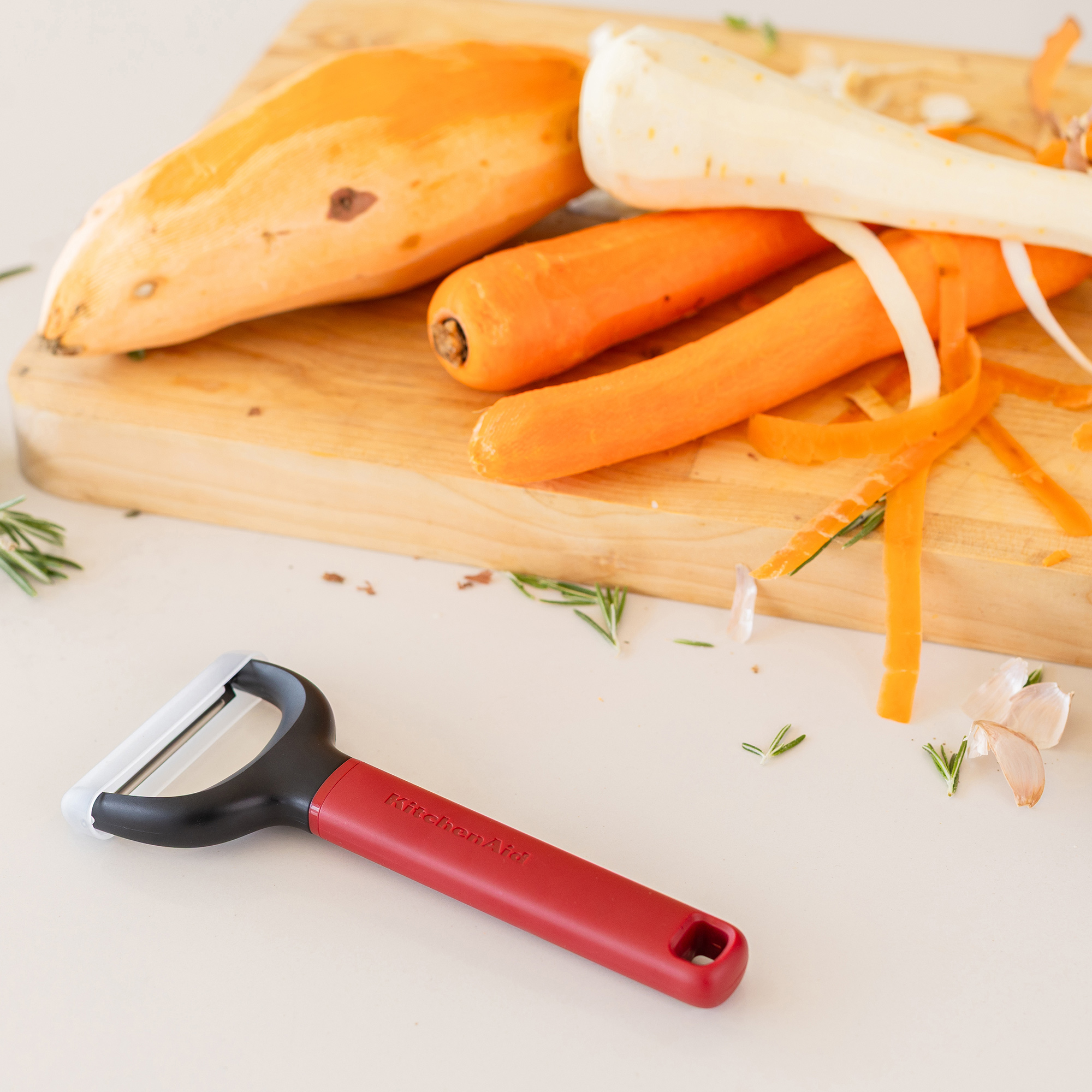 Ein roter Sparschäler liegt vor einem Holzbrett mit Karotten, Süßkartoffeln und Knoblauch.