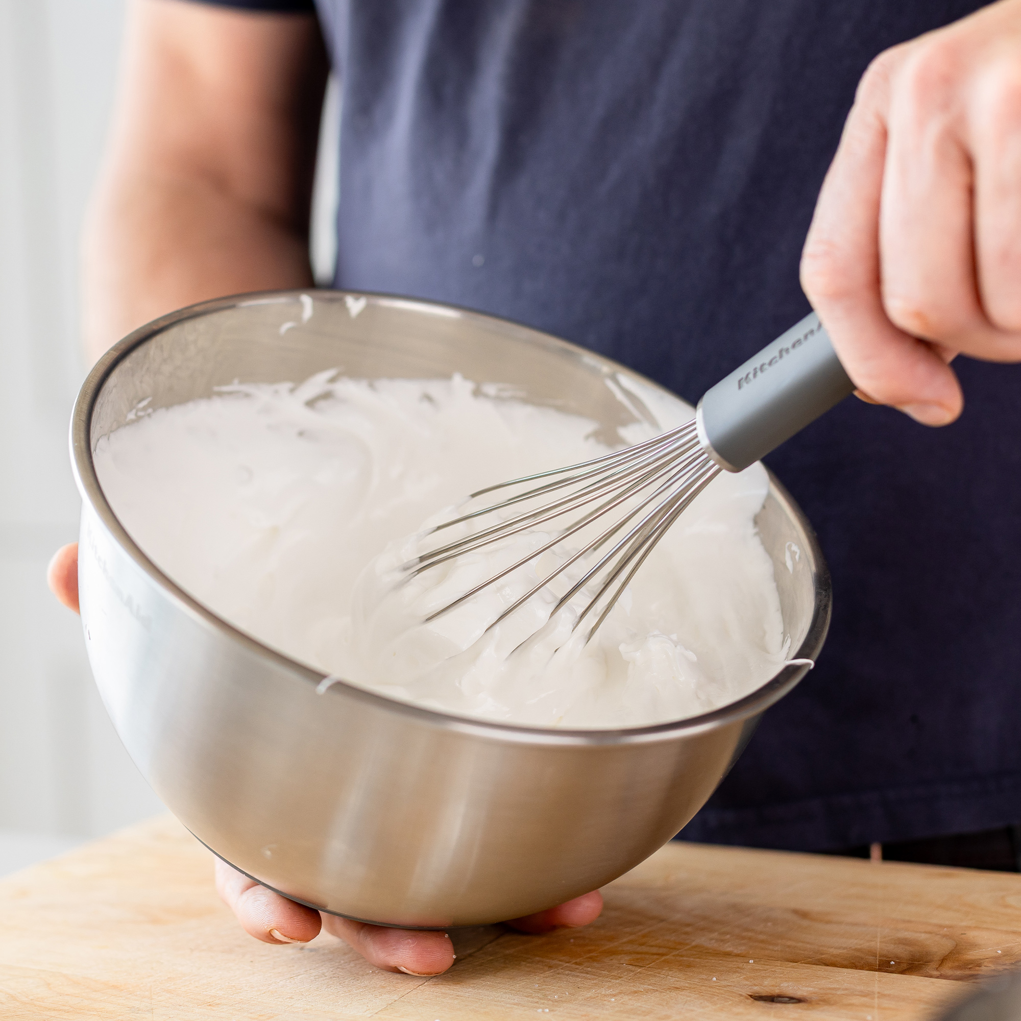 Eine Person schlägt weiße Creme in einer Edelstahlschüssel mit einem Schneebesen.