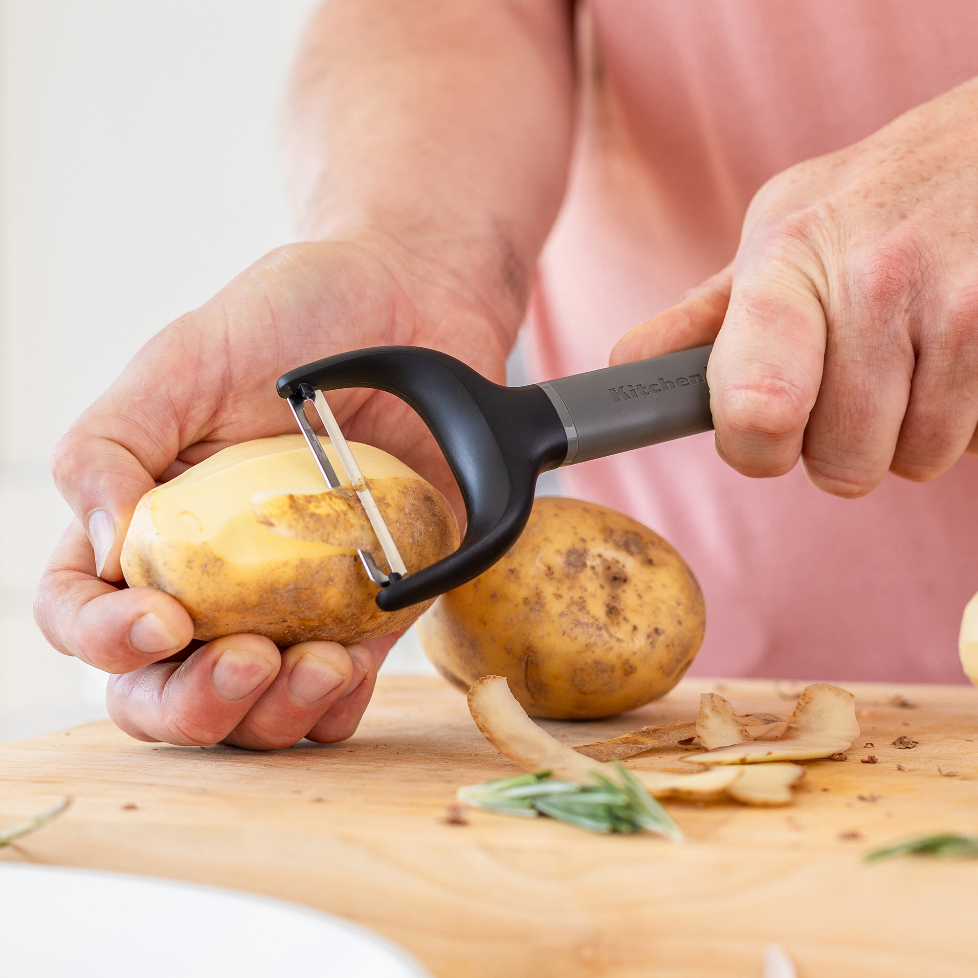 Hände schälen Kartoffeln mit einem Sparschäler auf einem Holzbrett mit Rosmarin.