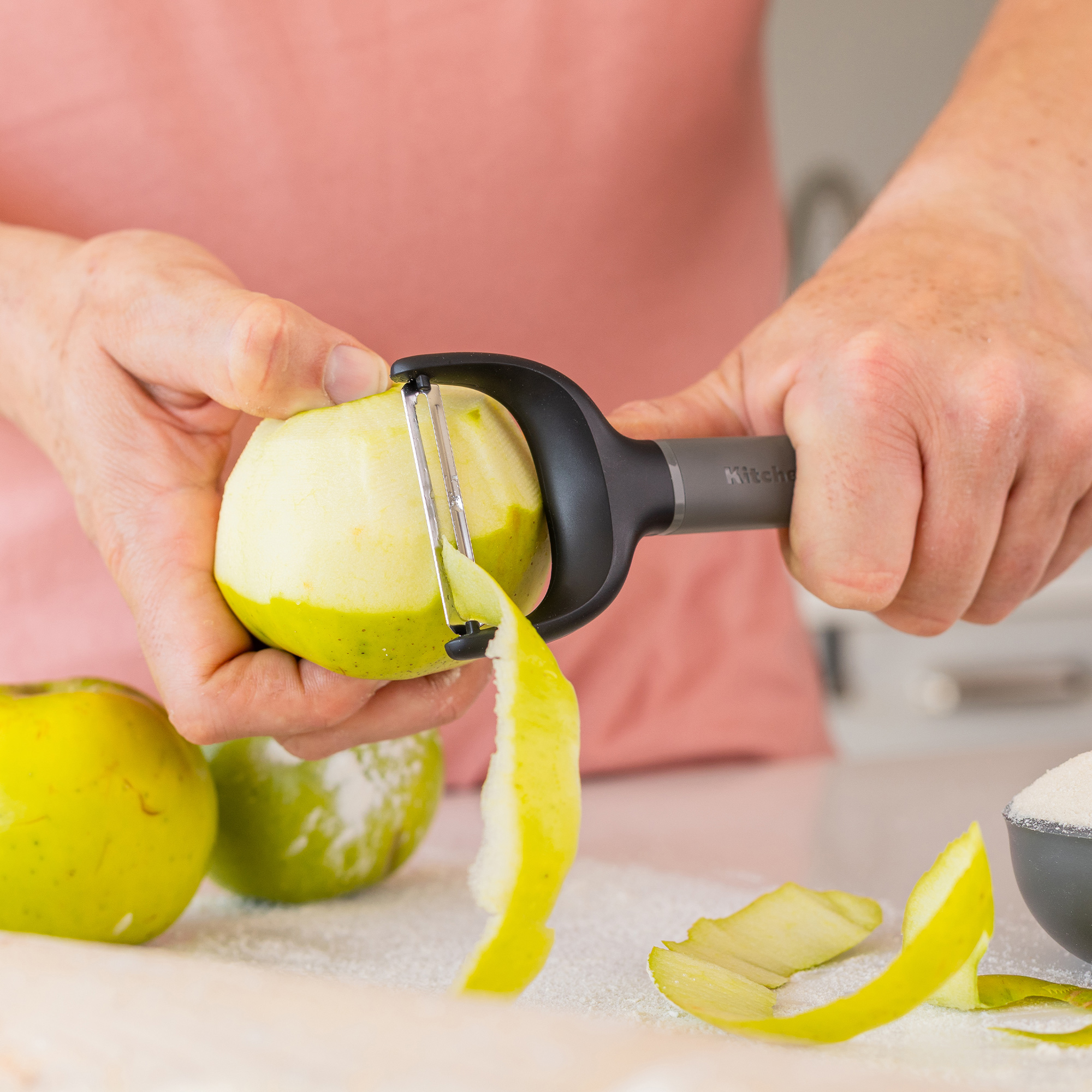 Hände schälen einen grünen Apfel mit einem Sparschäler, Apfelschale liegt daneben.