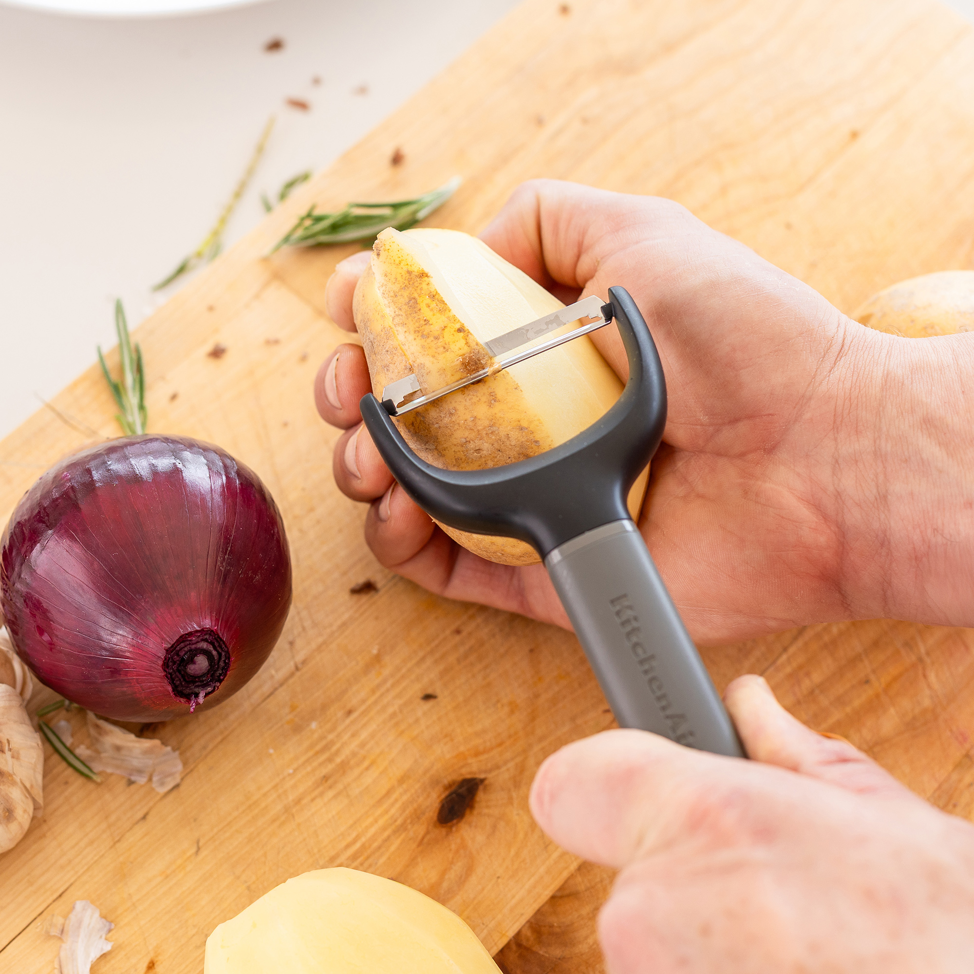 Hände schälen eine Kartoffel mit einem Sparschäler auf einem Holzbrett neben einer Zwiebel.