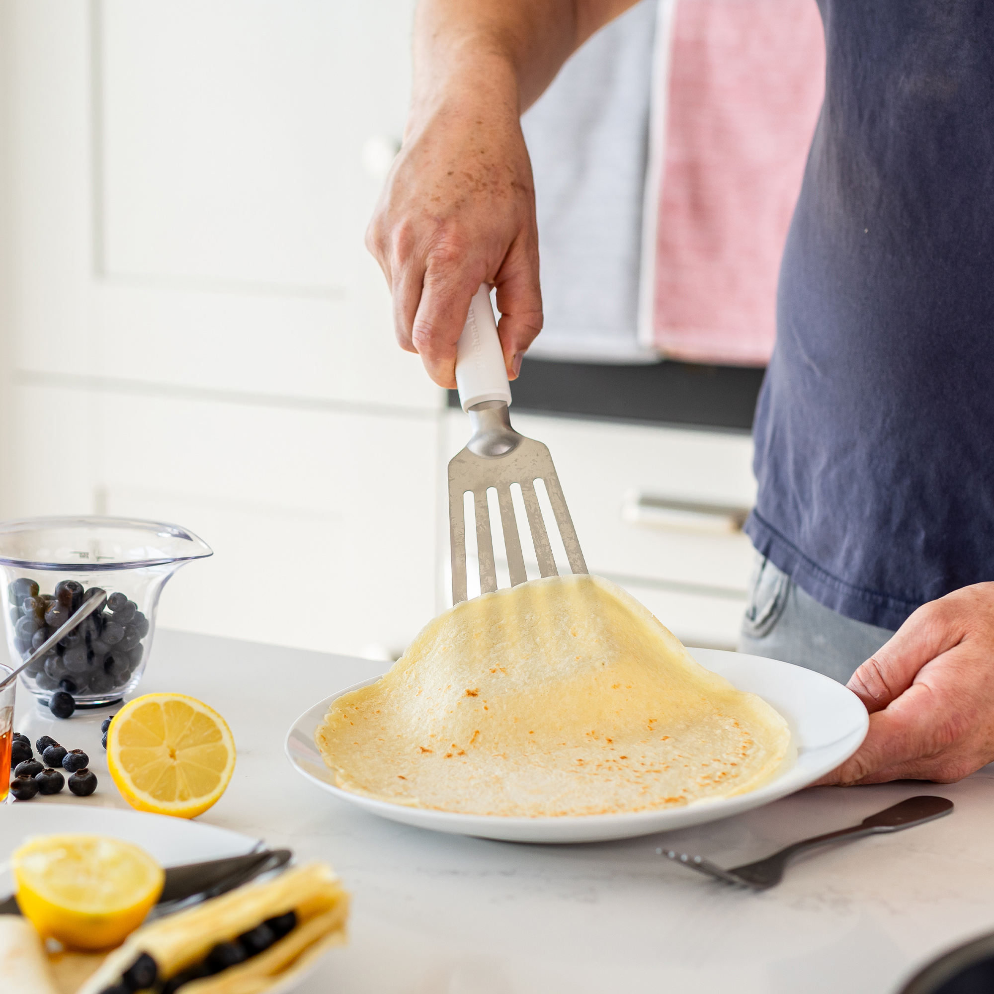 Eine Person faltet einen Crêpe mit einem Spatel auf einem Teller. Blaubeeren und Zitrone sind zu sehen.