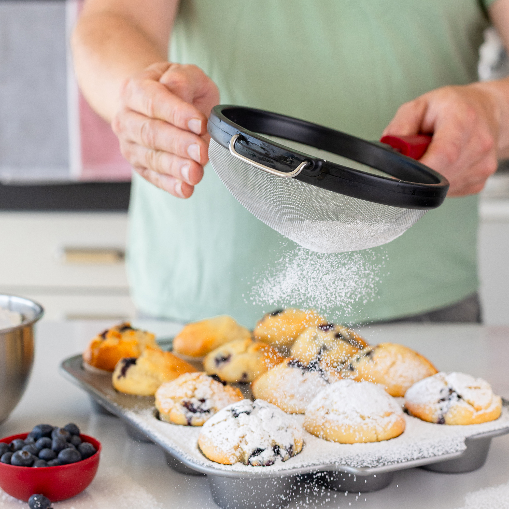 Puderzucker wird mit einem Sieb über Blaubeermuffins in einer Metallform gesiebt.