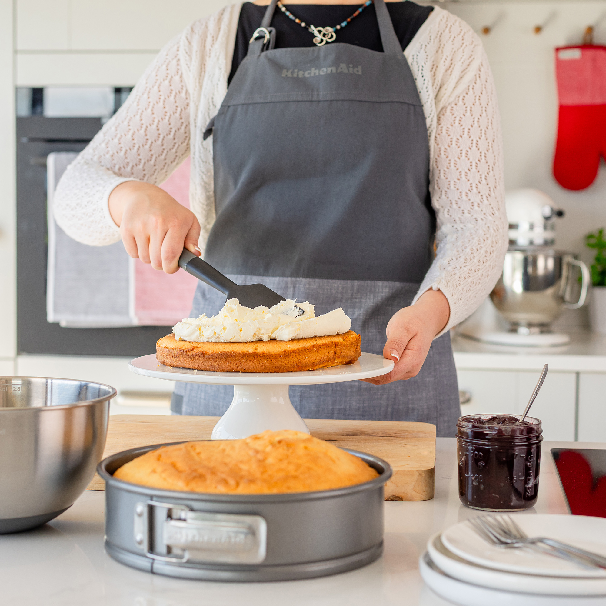 Eine Person verteilt Zuckerguss auf einem Kuchen. Eine Rührschüssel, eine Kuchenform und ein Marmeladenglas sind in der Nähe.