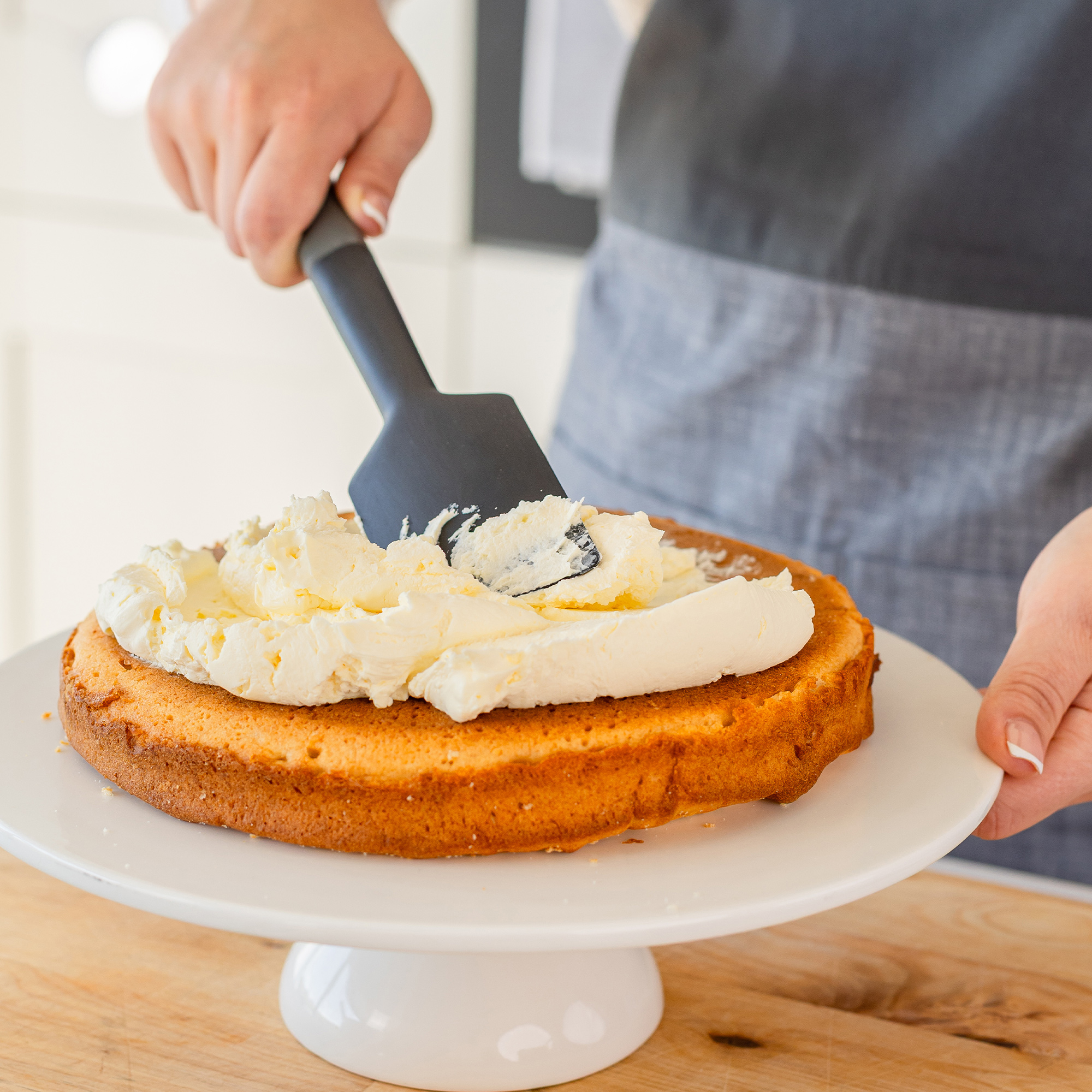 Eine Hand verteilt weiße Creme auf einem Kuchenboden auf einem weißen Tortenständer.