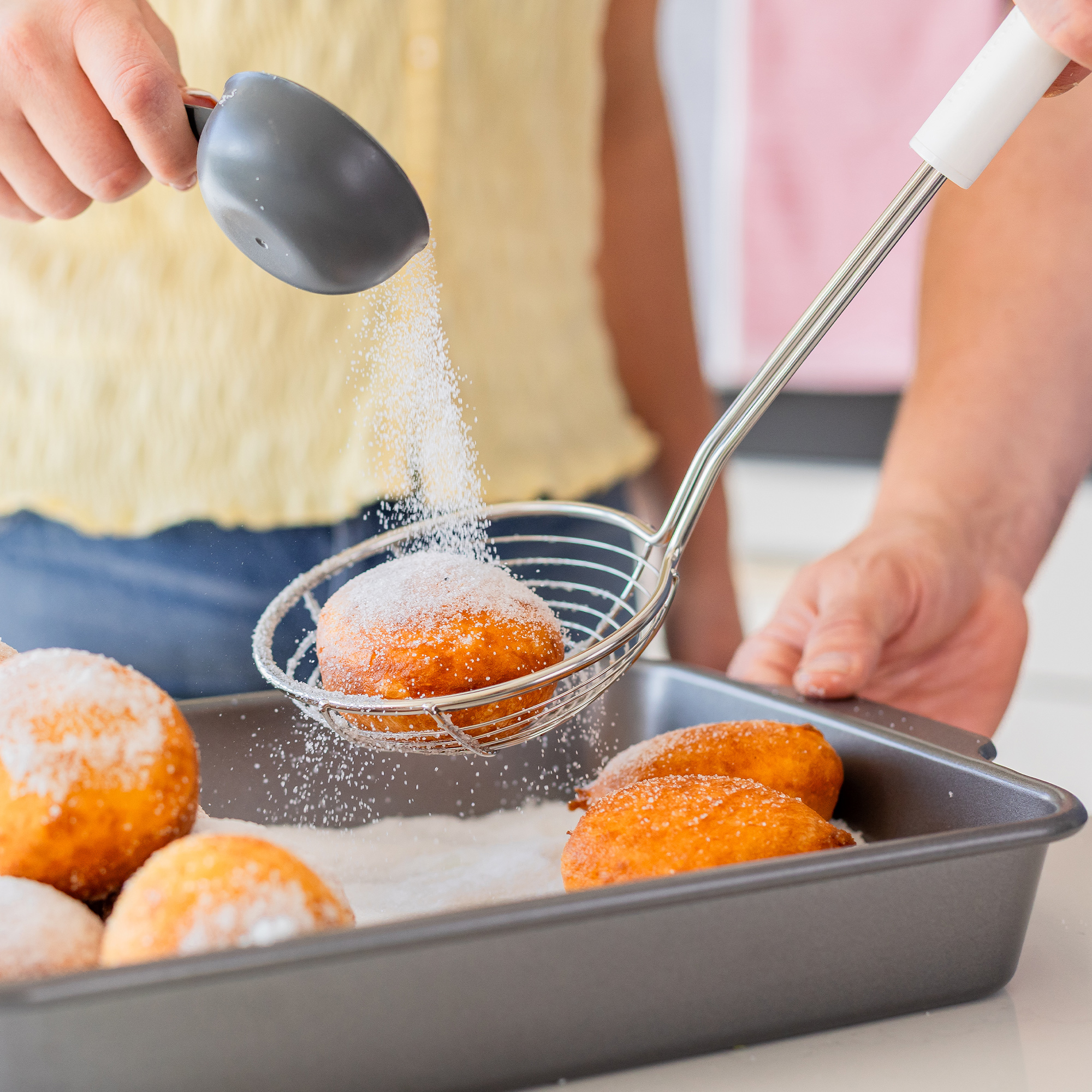 Zucker wird auf goldbraune Donuts gestreut, die in einem Metallsieb über einem Backblech gehalten werden.