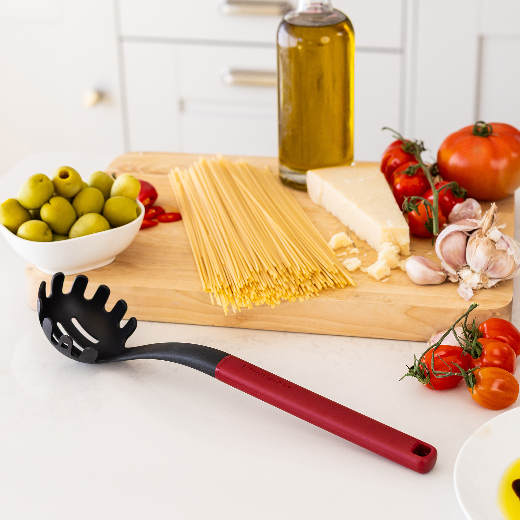 Spaghetti, Oliven, Tomaten, Knoblauch, Parmesan, Öl und Nudellöffel auf einem Holzbrett.