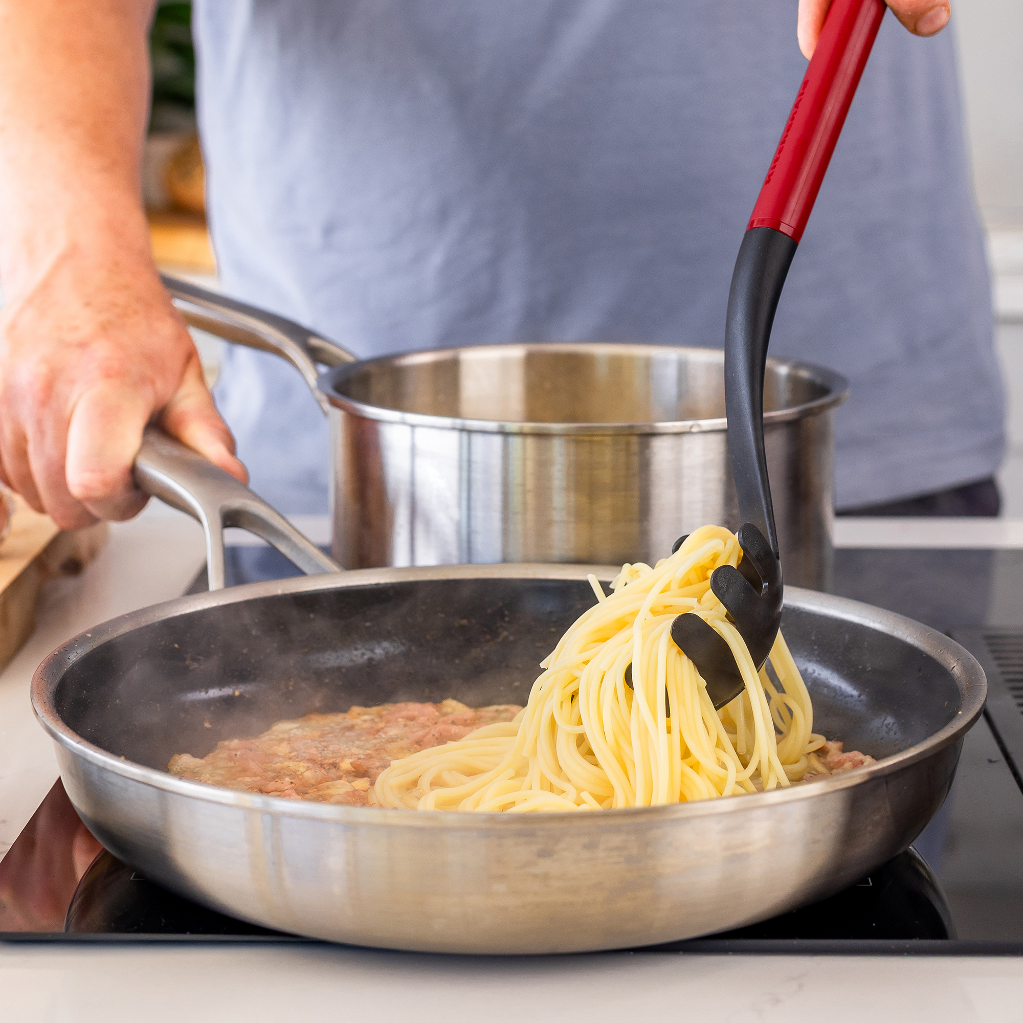 Spaghetti werden mit einer Zange in eine Pfanne mit Speck gegeben.