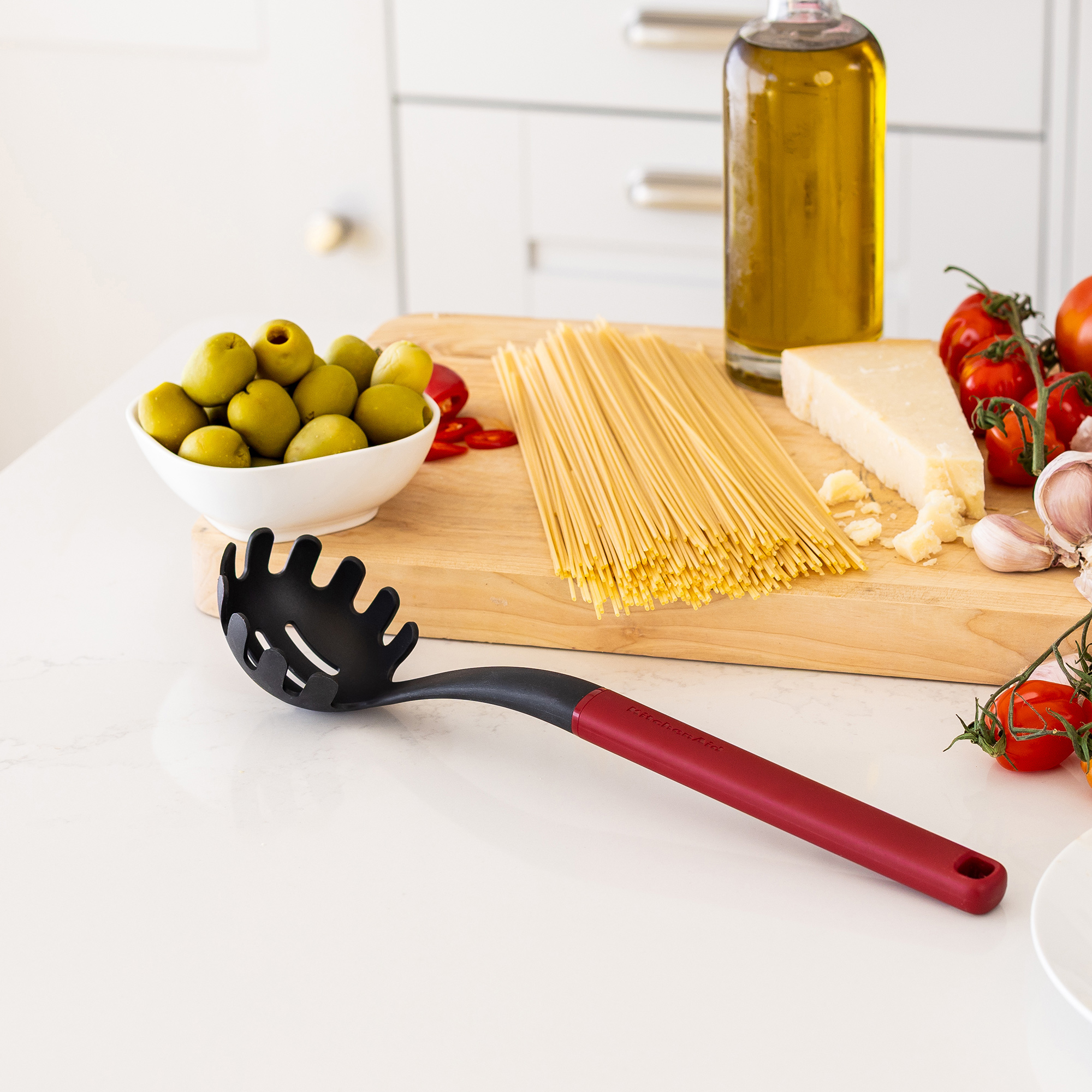 Ein Nudellöffel liegt vor Zutaten für Pasta: Spaghetti, Oliven, Tomaten, Käse, Knoblauch, Öl.
