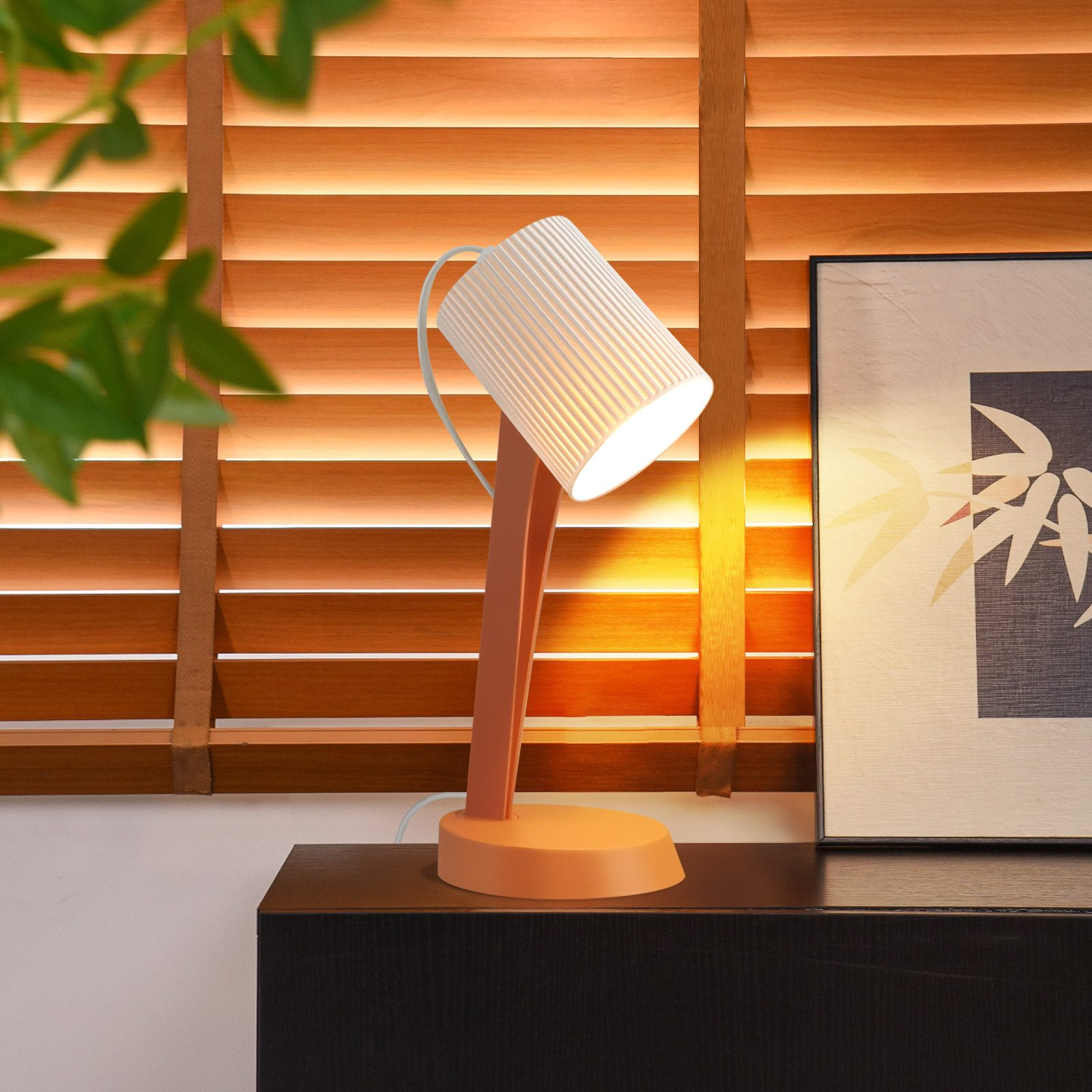 Una moderna lampada da scrivania con paralume bianco a coste è appoggiata su un mobile scuro, vicino a veneziane in legno e un quadro.