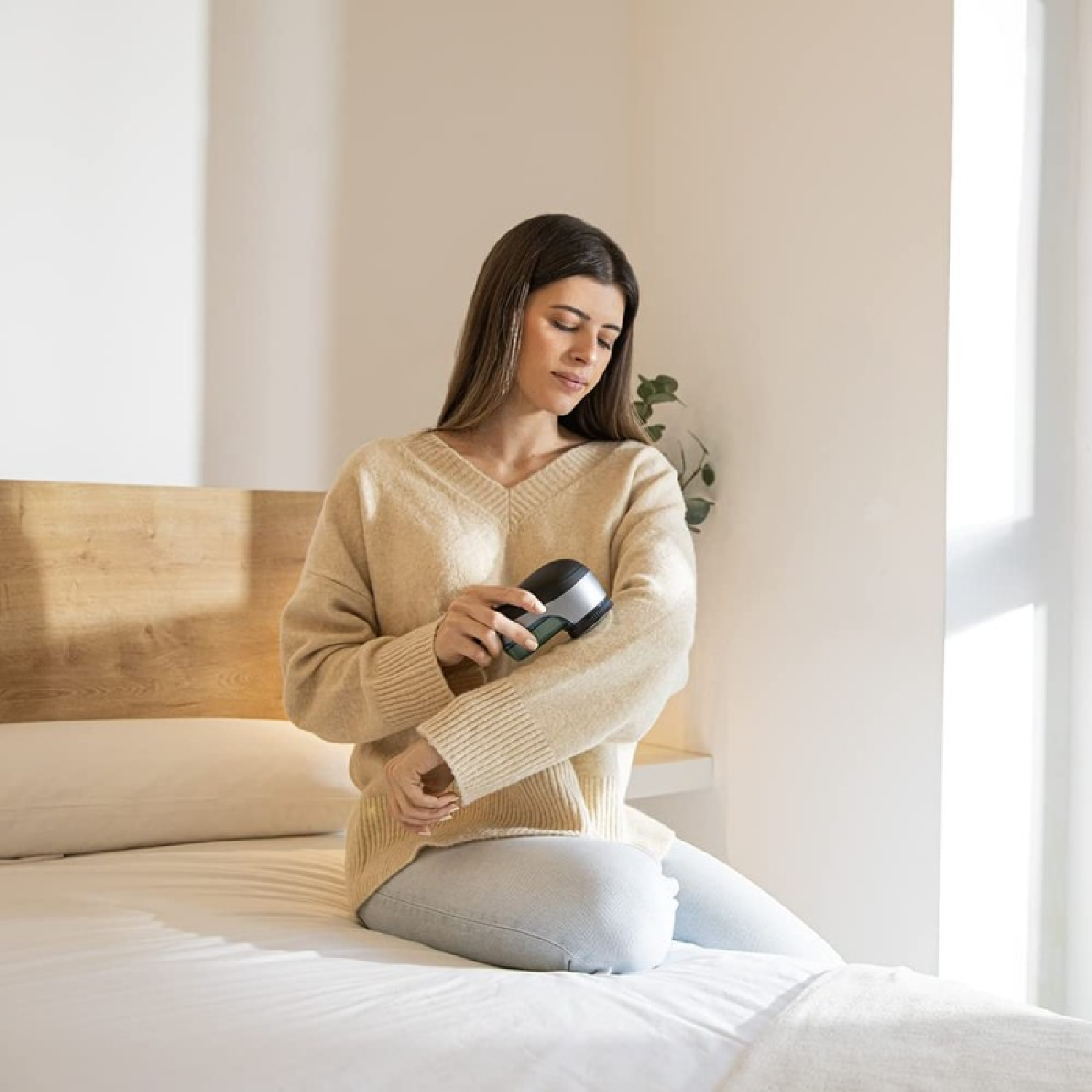 Una donna con un maglione beige è seduta su un letto e usa un levapelucchi grigio scuro e argento sulla manica.