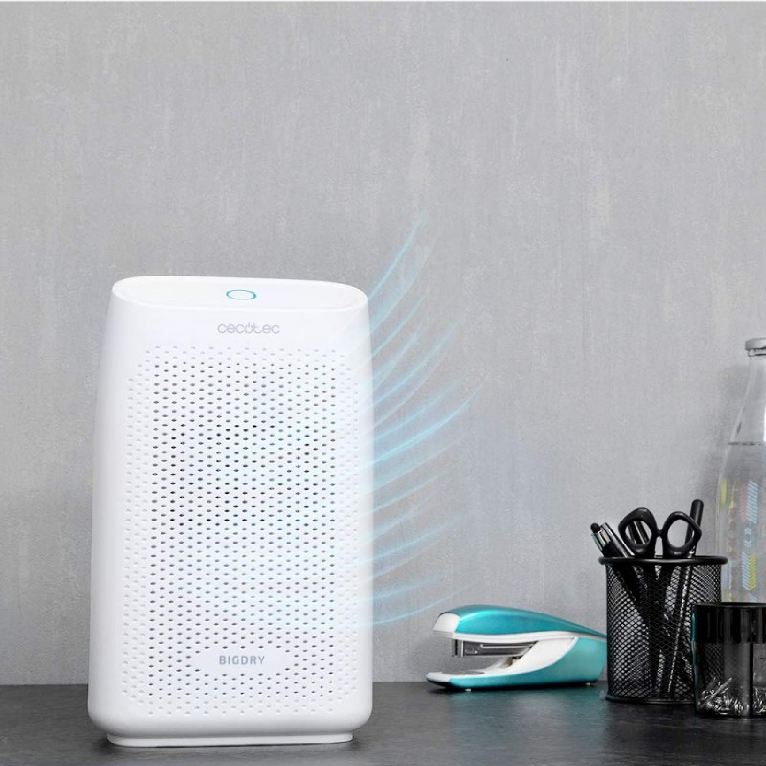 Biały osuszacz powietrza stoi na stole obok materiałów biurowych i butelki.