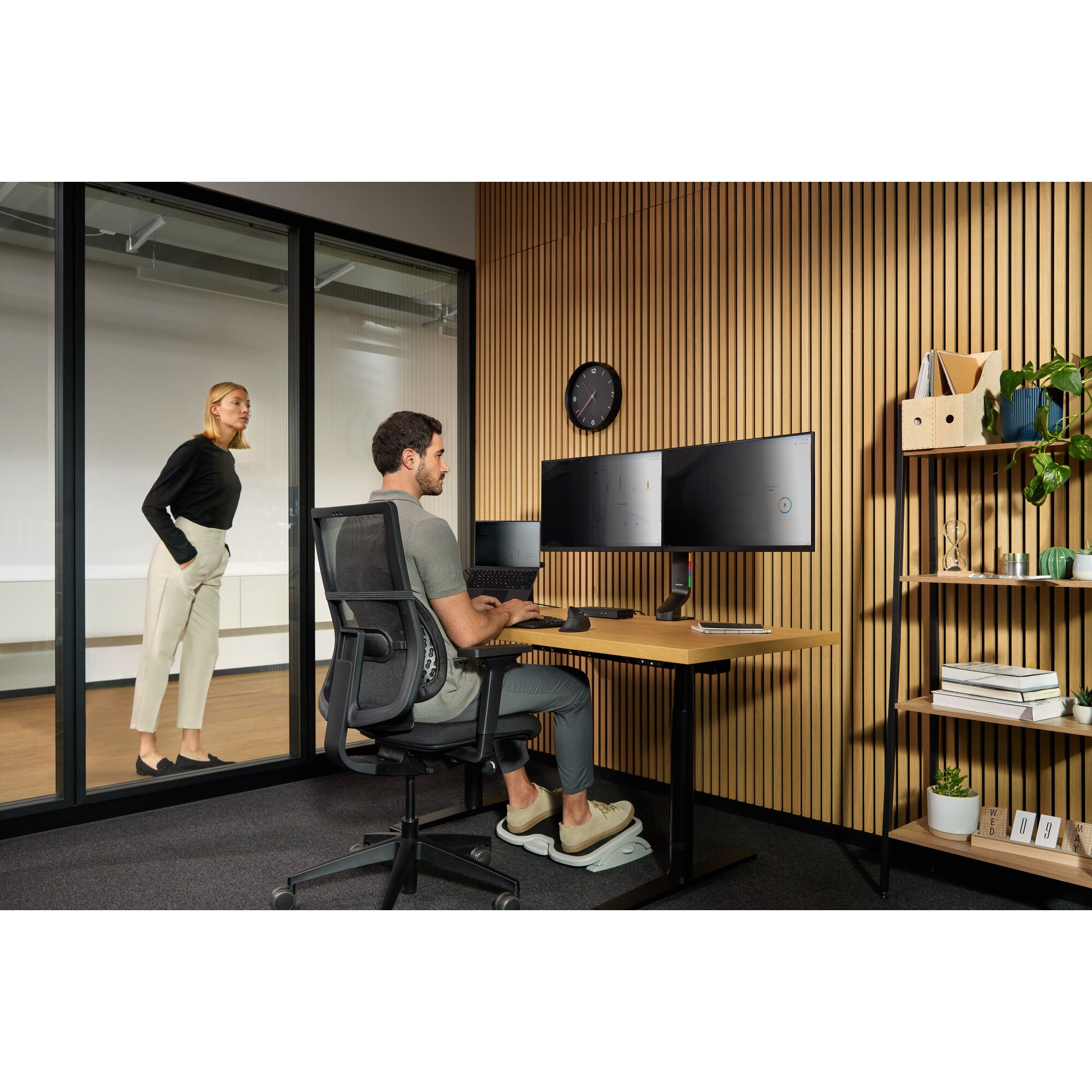 Mann am Schreibtisch mit zwei Monitoren, Frau im Glasbüro. Holzwand.