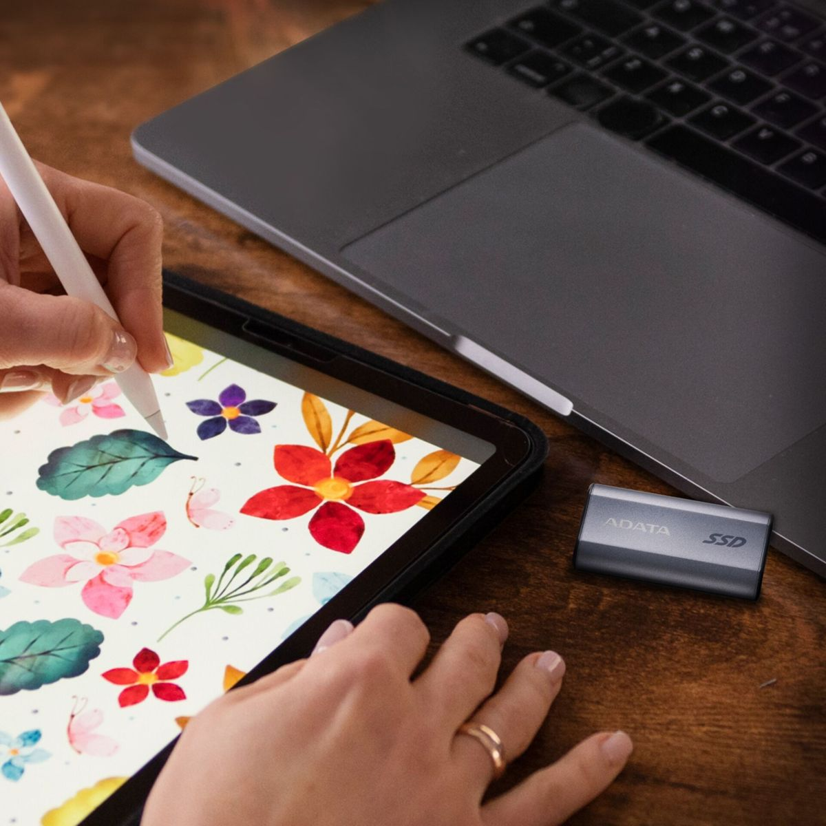 Une main dessine sur une tablette à motif floral à côté d'un ordinateur portable et d'un SSD.