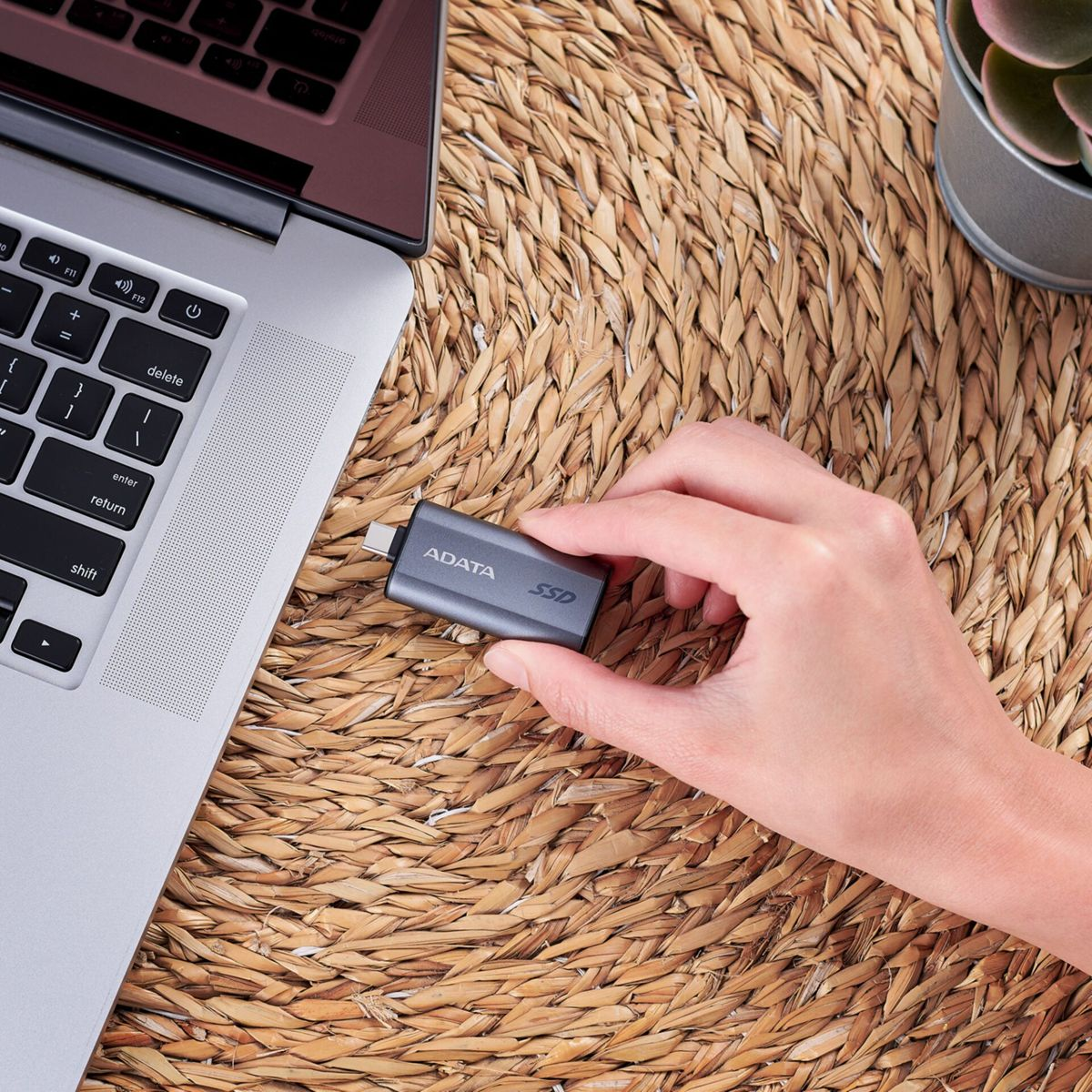 Une main tient un SSD ADATA gris à côté d'un ordinateur portable sur une surface tressée.