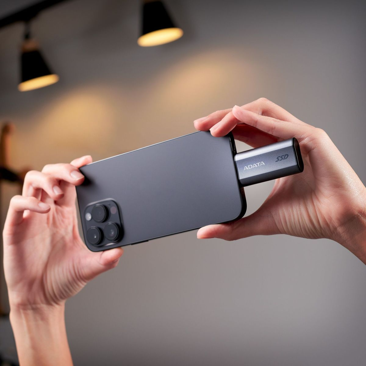 Des mains tiennent un smartphone gris foncé. Un SSD ADATA est branché au téléphone.