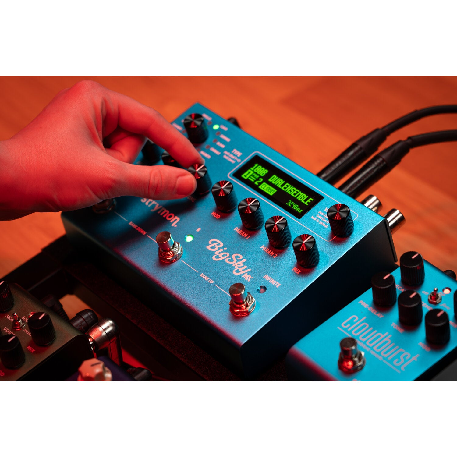 Een hand stelt een blauw Strymon Big Sky reverb pedaal af op een pedalboard met andere pedalen en kabels.