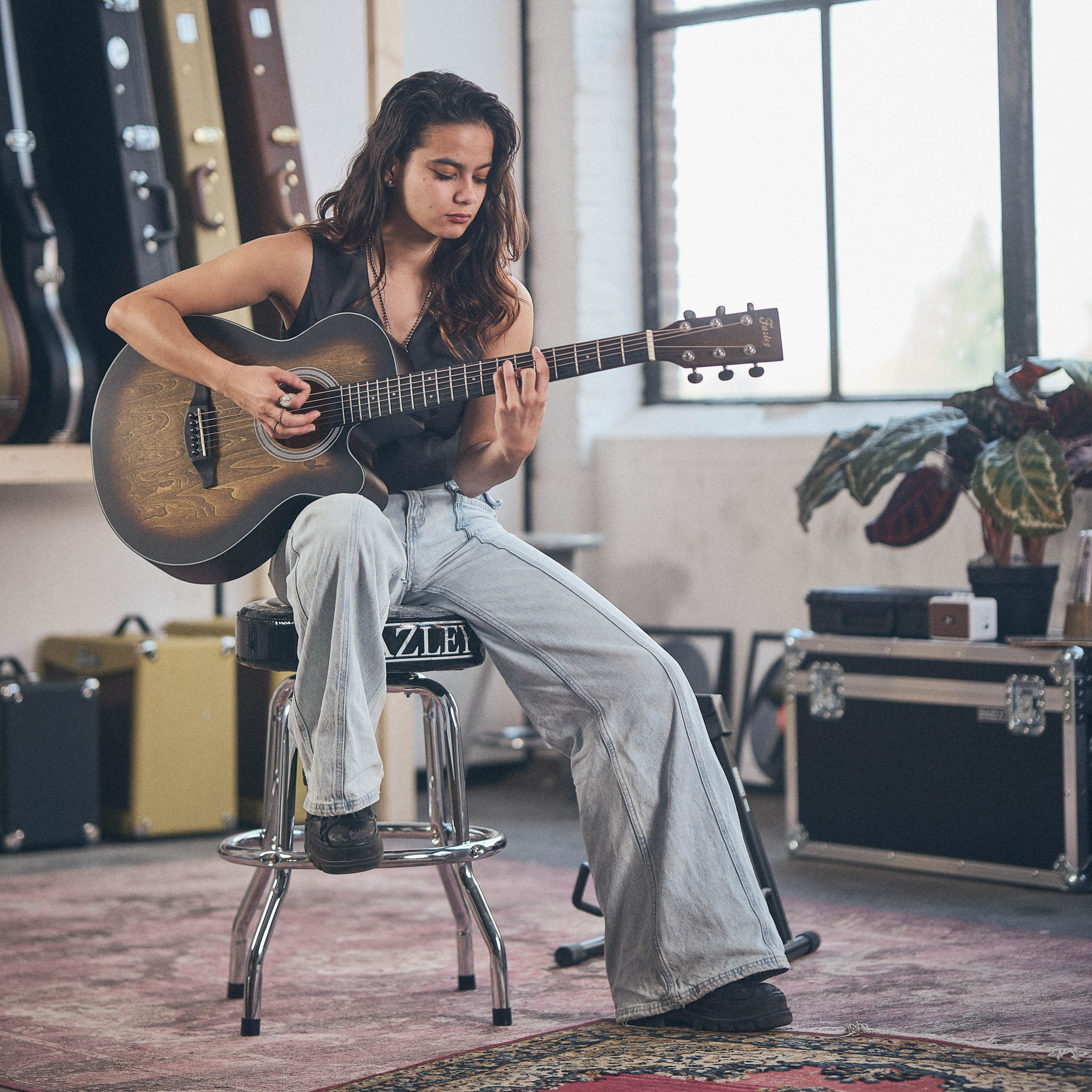 Een vrouw zit op een kruk en speelt gitaar in een kamer met gitaarkoffers.