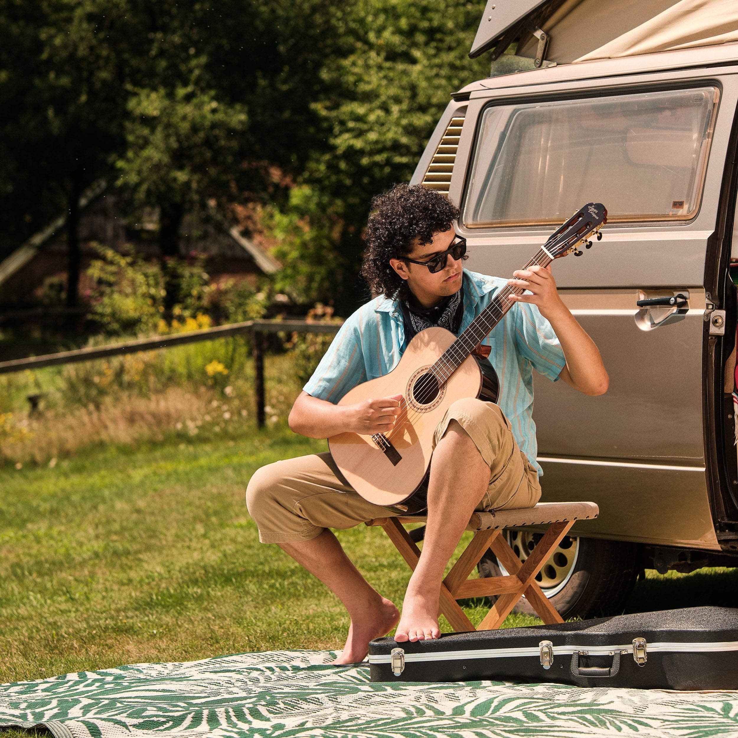 Een man met krullend haar speelt gitaar naast een camper op een grasveld.