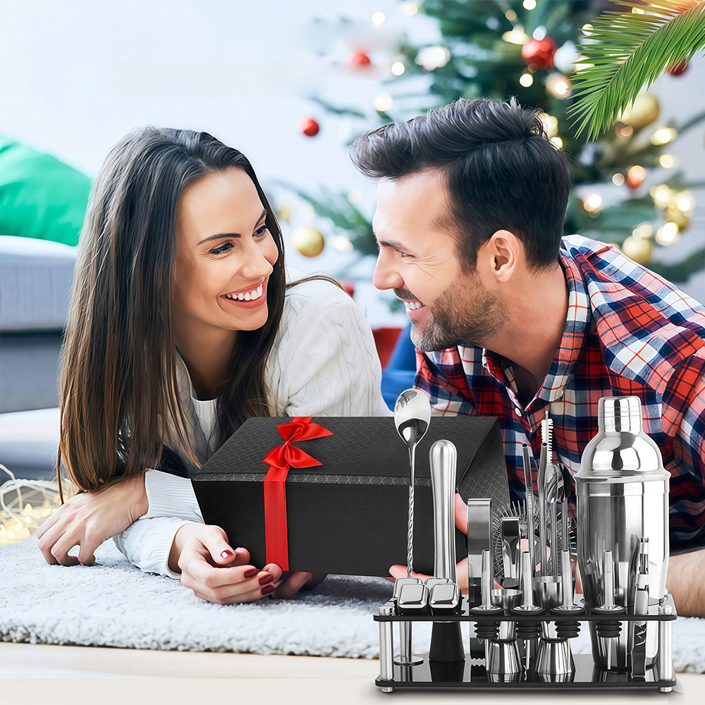 Ein Paar liegt auf einem Teppich vor einem Weihnachtsbaum, umgeben von einem Geschenk und Cocktail-Werkzeugen.