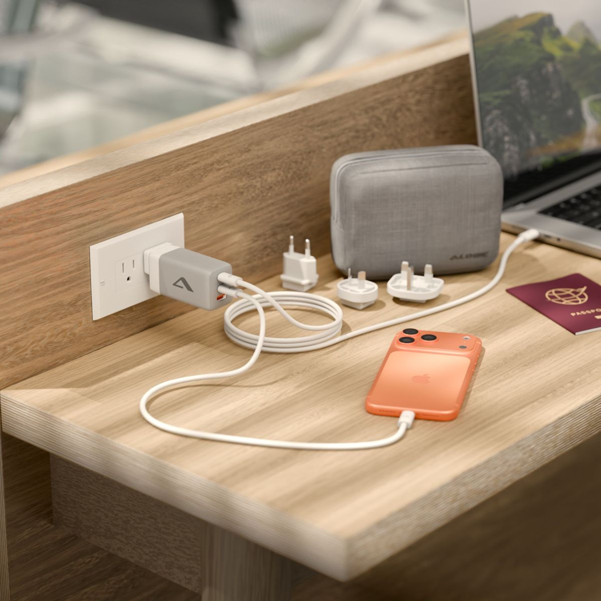 Telefono arancione si carica accanto a laptop, passaporto e adattatori da viaggio sulla scrivania.