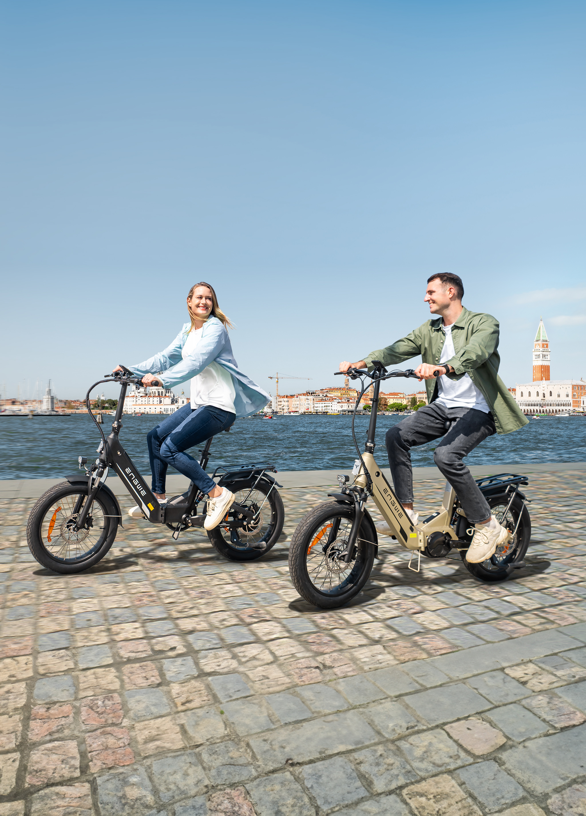 Ein Mann und eine Frau fahren auf E-Bikes auf einem gepflasterten Weg am Wasser.