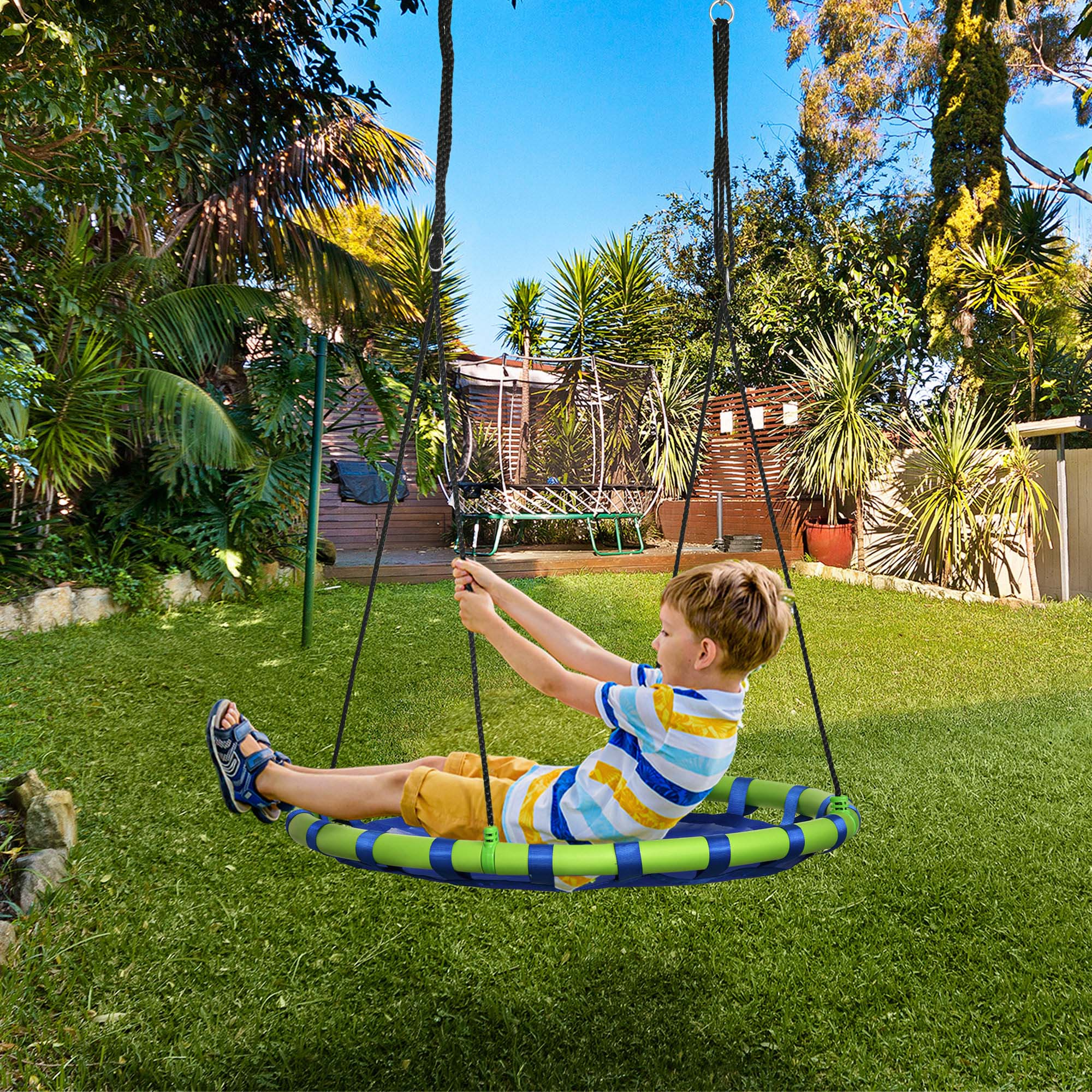 Un niño se columpia en un columpio redondo en un patio verde con árboles y trampolín.