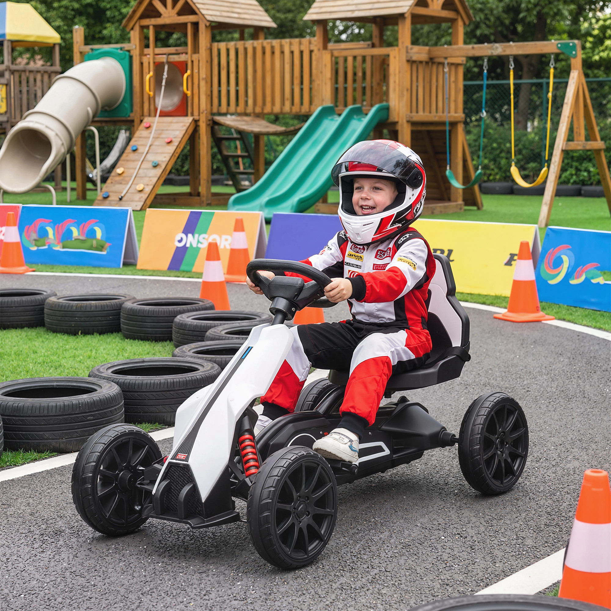 Un niño con traje de carreras conduce un go-kart en una pista con parque infantil detrás.