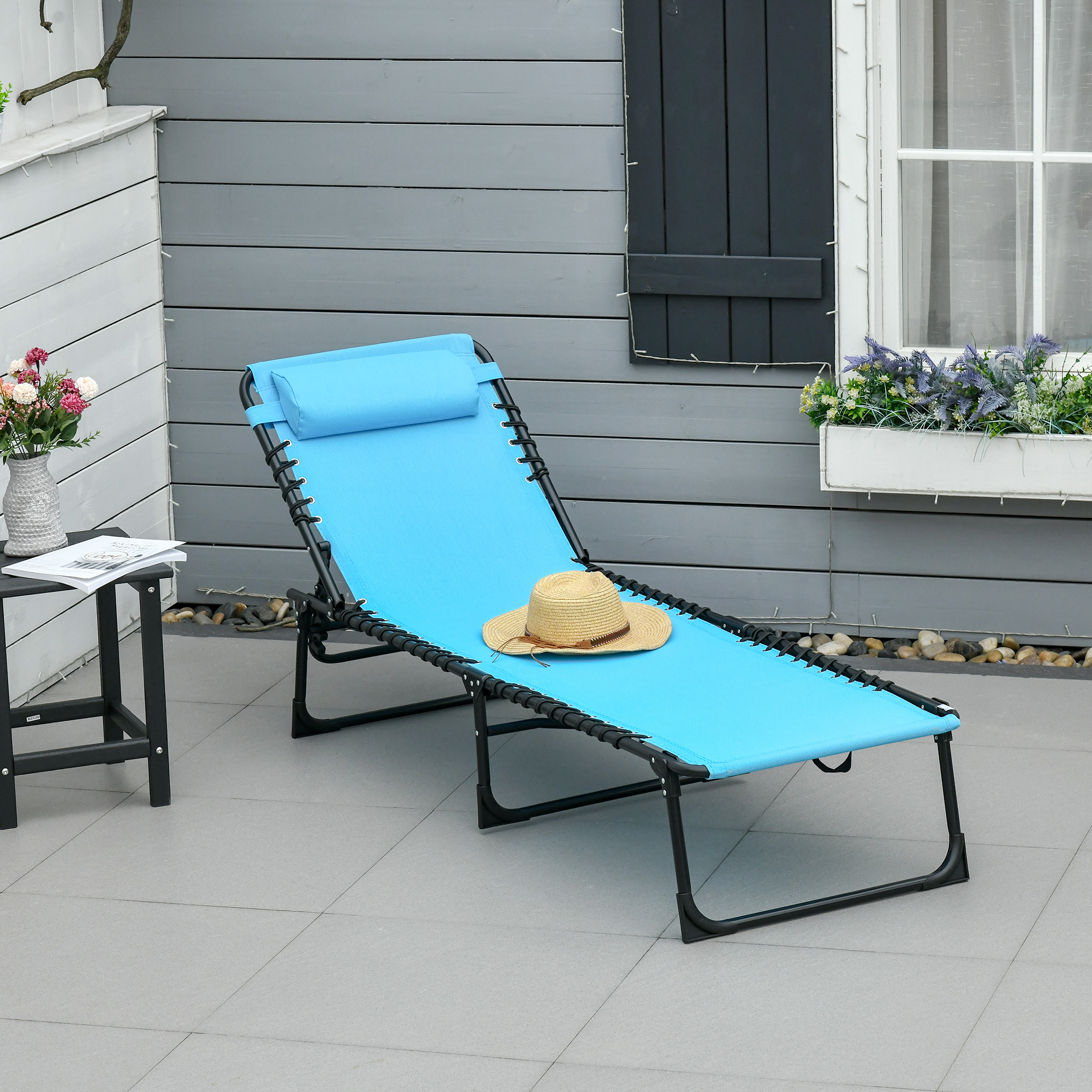 Una tumbona azul con cojín y sombrero está en un patio junto a una mesa con flores.