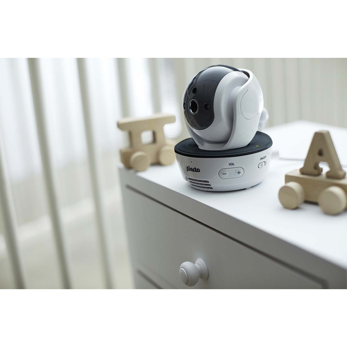 Ein weißer Babyfon mit schwarz-grauem Kopf steht auf einer weißen Kommode neben Holzspielzeug.