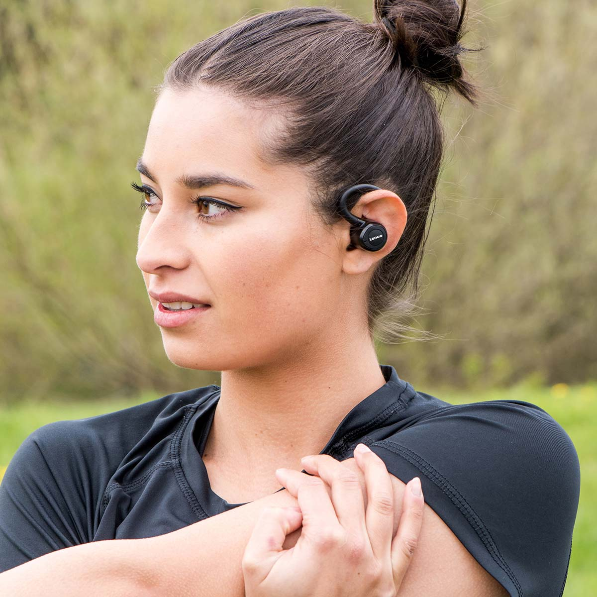 Mujer con camiseta negra y auriculares negros, estirándose al aire libre con fondo verde borroso.